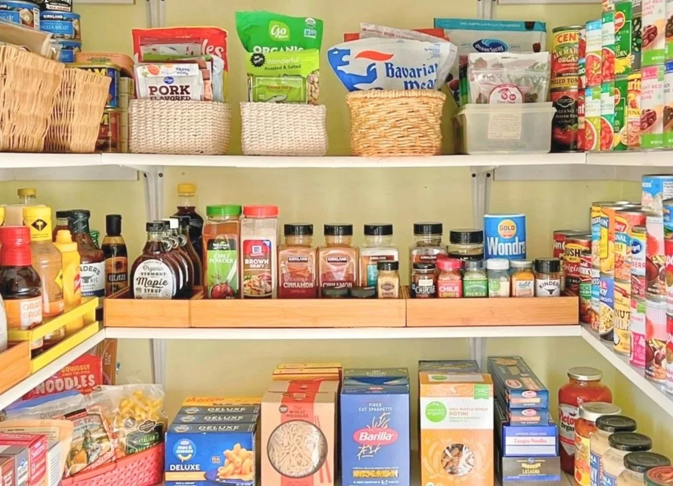 Organized pantry shelf with baskets containing snack foods, spices, sauces, and canned goods, including noodles, syrups, and canned vegetables.