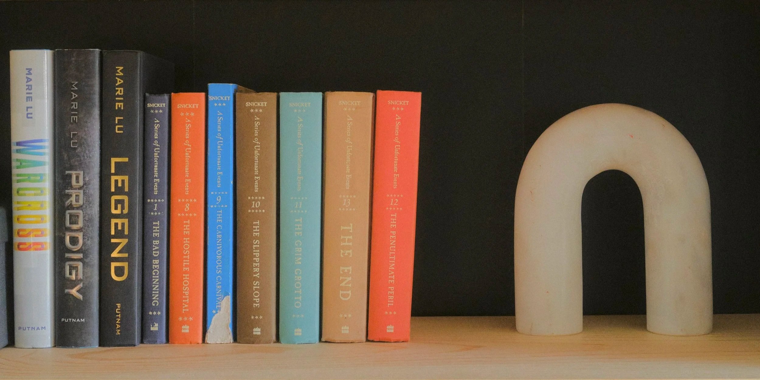 A shelf with books and a rectangular arch shaped object, all placed on a light-colored wooden surface.