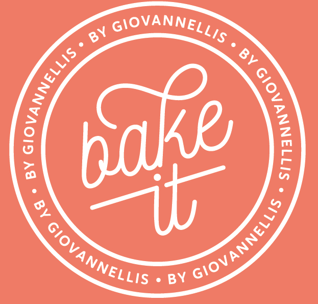 A circular logo with a coral background and white text. The inner text says 'bake it' in a cursive font. The outer ring features the text 'BY GIOVANNELLIS' repeated four times, separated by small dots.