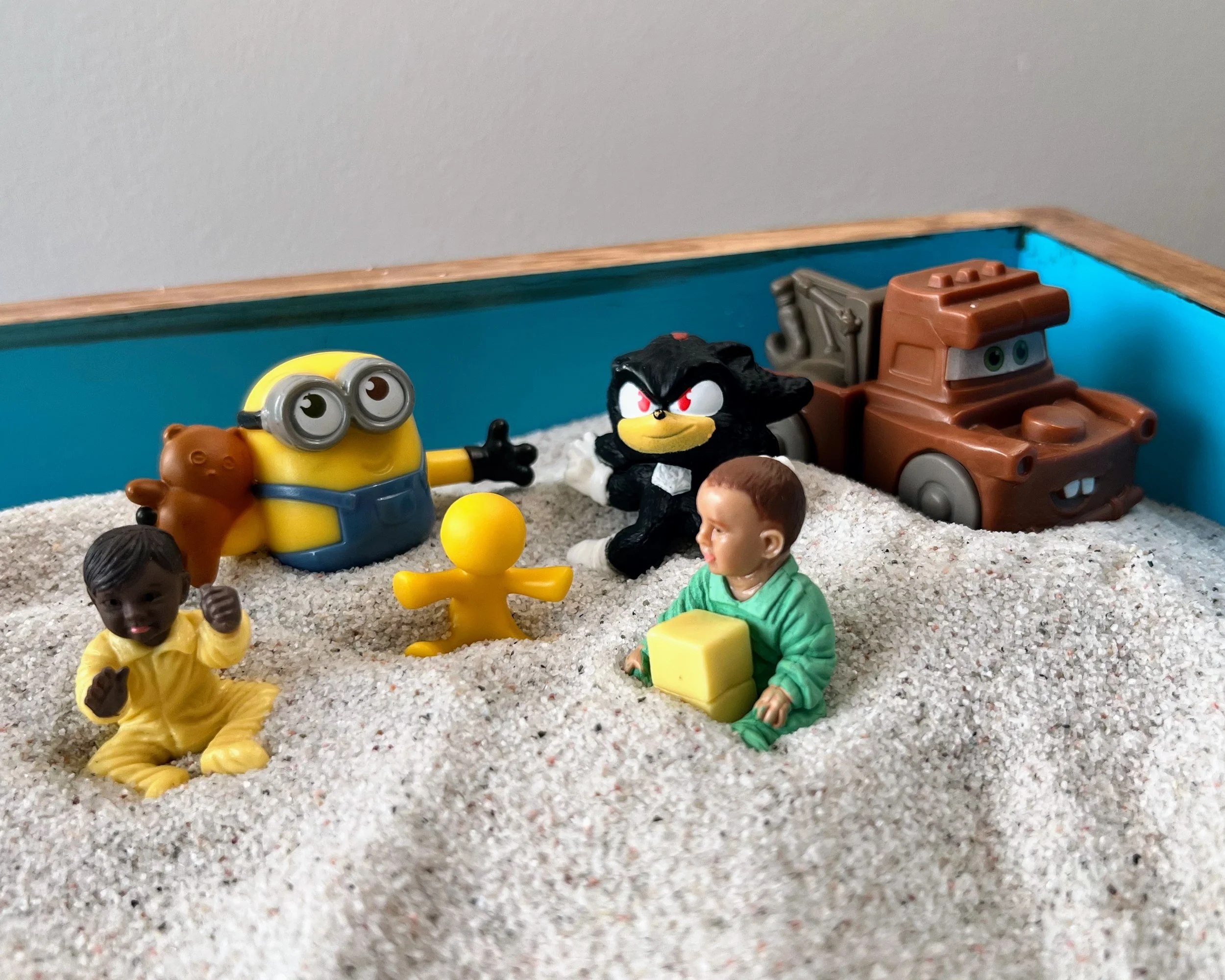Image shows a corner of the sand tray with white sand and blue edges. Inside are miniatures of Minions, Shadow the Hedgehog, Mater, and three infants of different skin tones.