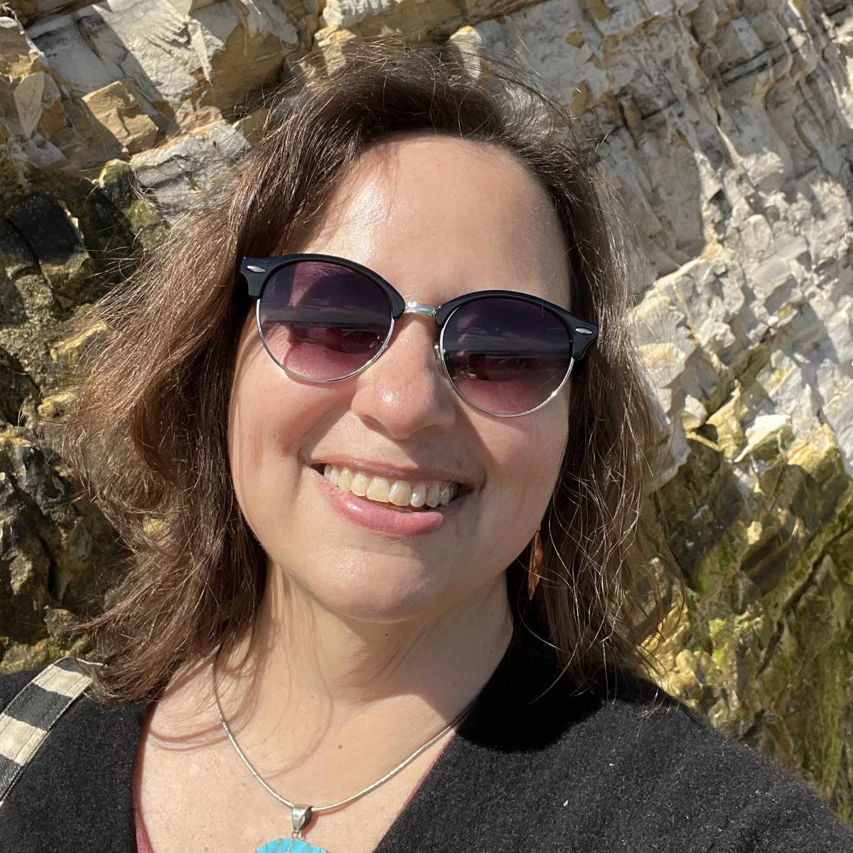 Picture shows a woman with fair skin, long brown hair wearing Polaroid glasses and standing in front of a cliff face.