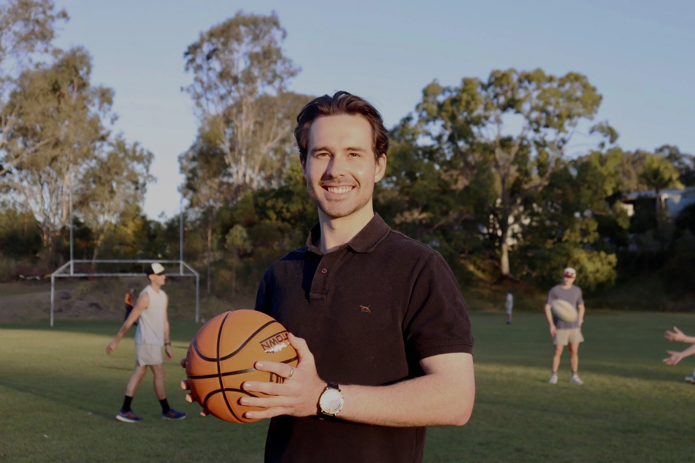 A young man holding a basketball on a grassy field with other people playing rugby in the background, trees, and a clear sky.