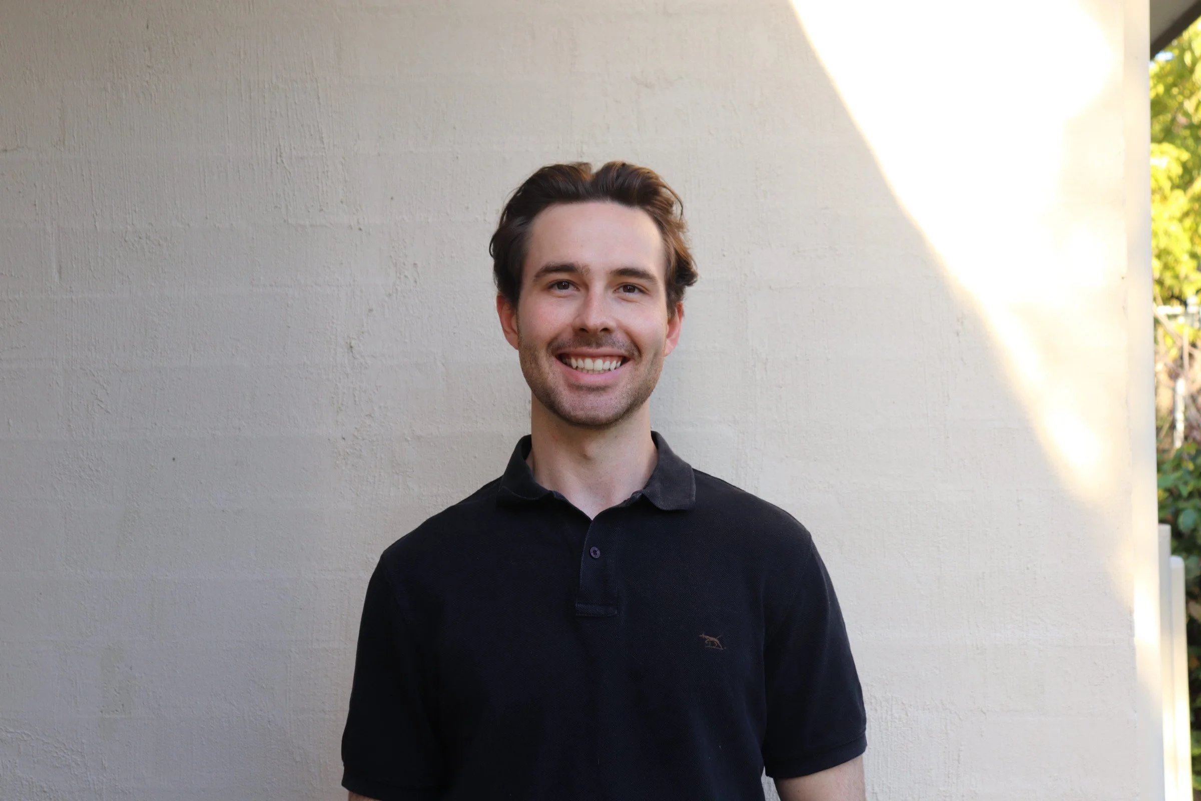 A young man with brown hair and a beard smiling, standing outdoors against a light-colored wall with sunlight and greenery in the background.