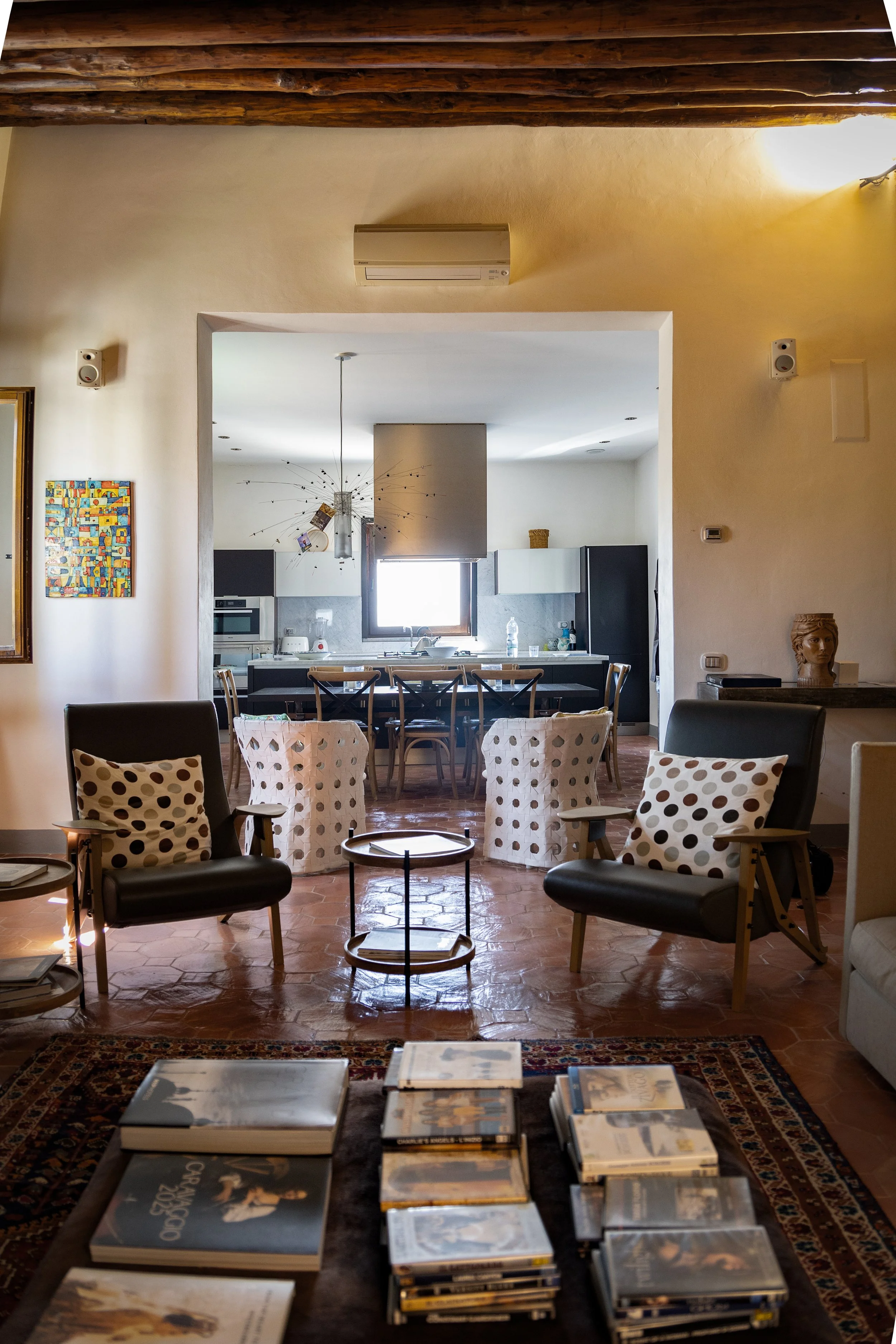 Living room with two black armchairs with polka dot pillows, a coffee table, and stacks of magazines. The background shows a dining area and kitchen with a window, dining table, and modern appliances.