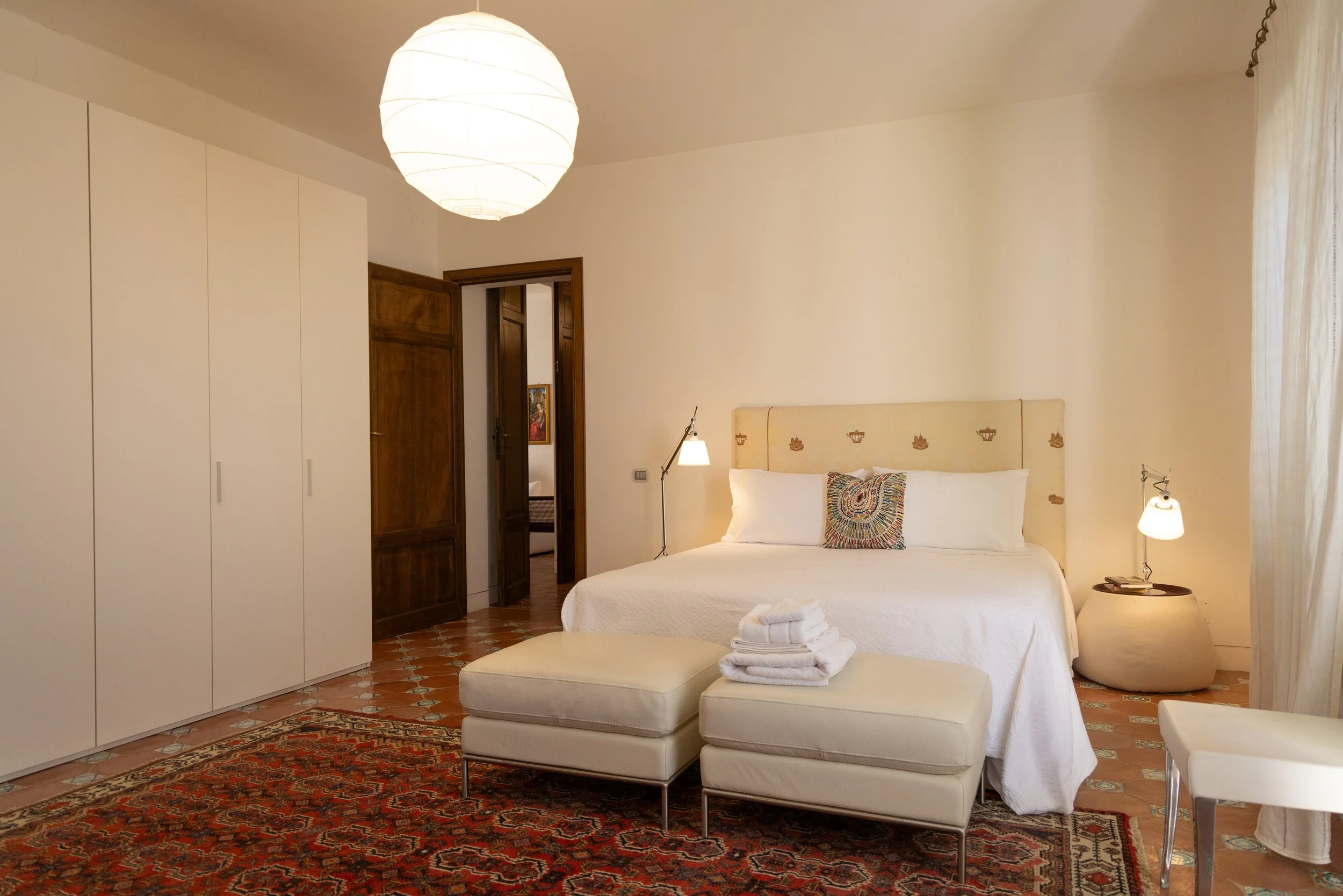 A bedroom with a white bed, colorful pillow, folded towels, and two white ottomans, with a large wardrobe and wooden door, terracotta tile floor, and a large paper lantern ceiling light.