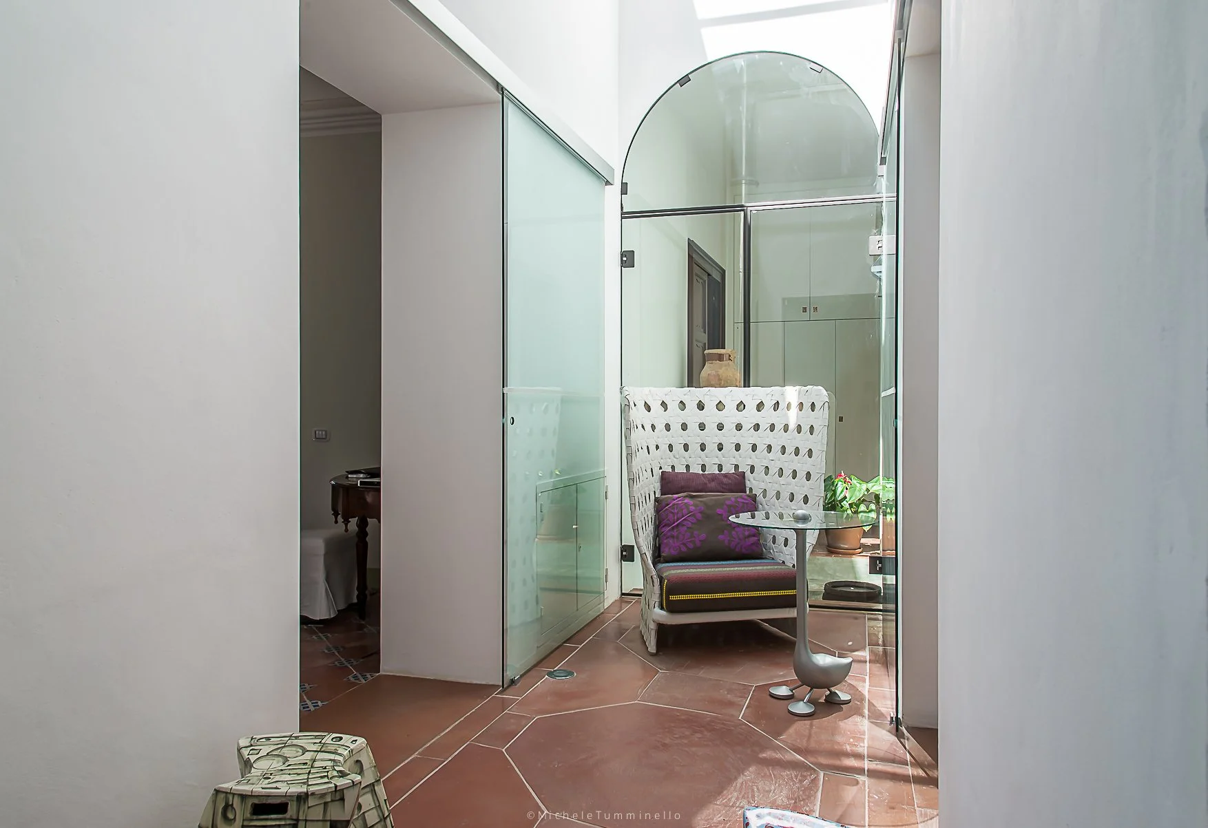 A cozy corner with a woven chair featuring a purple pillow and a small round glass table, located beside a window with potted plants, enclosed by glass walls and a high ceiling.