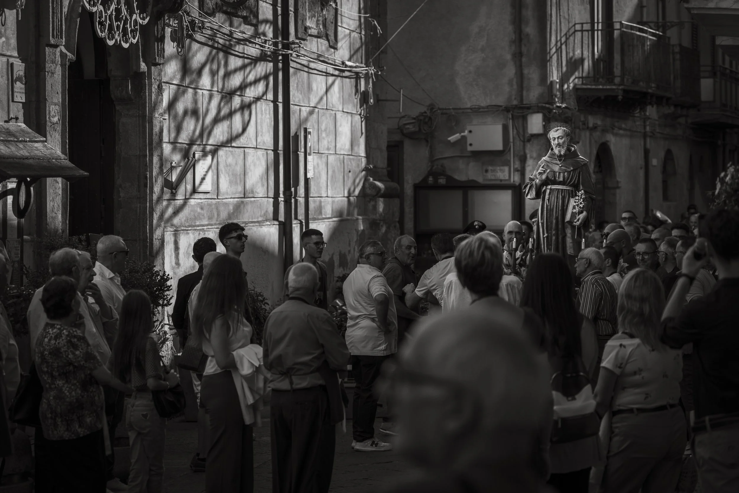 A black and white photo of a crowd gathered around a statue of a person in religious robes on a city street.