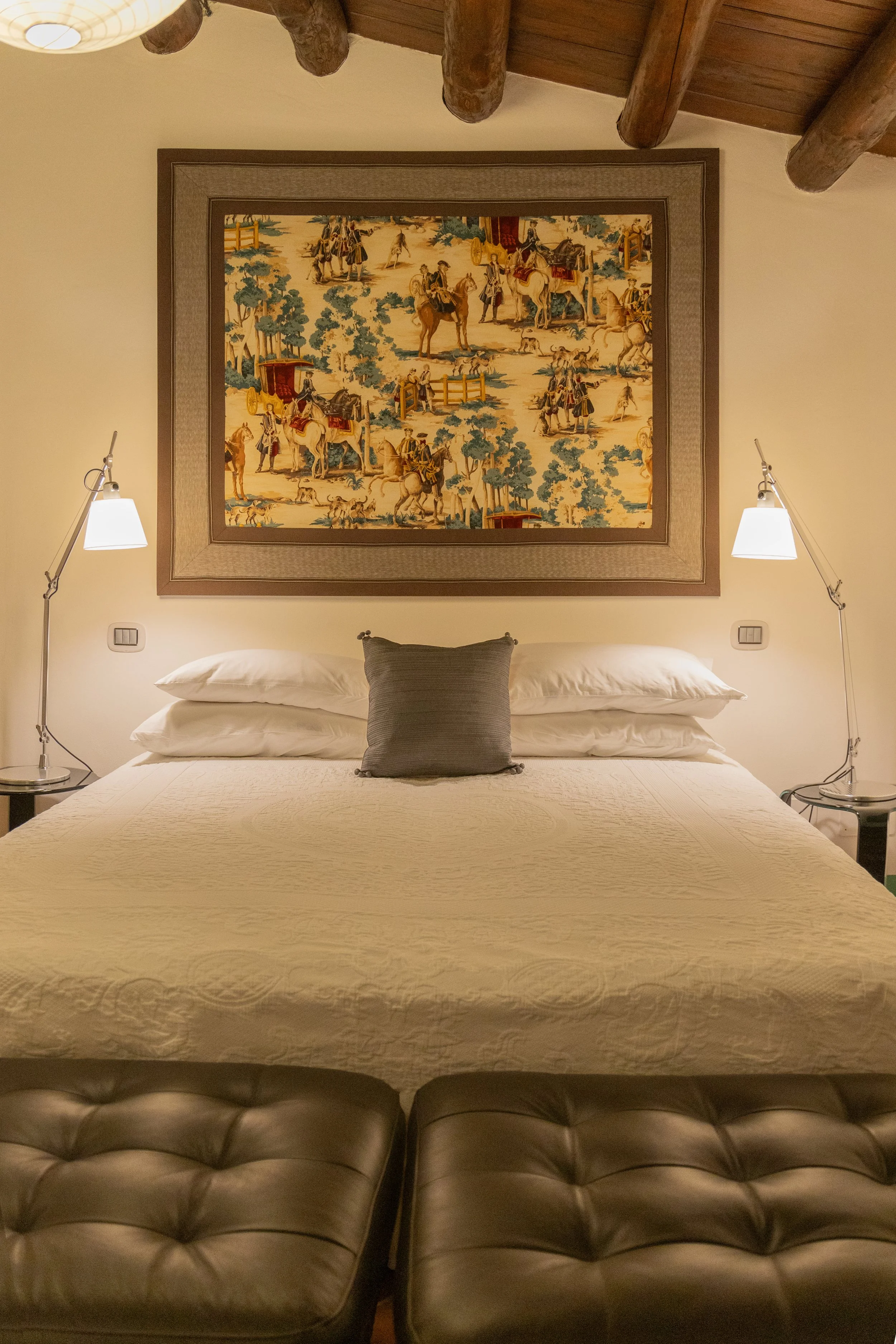 A neatly made bed with white bedding and a gray pillow in a cozy bedroom. Above the bed is a large framed painting depicting a scene with people, horses, and trees. There are two wall-mounted reading lamps on either side of the bed, and the room has 