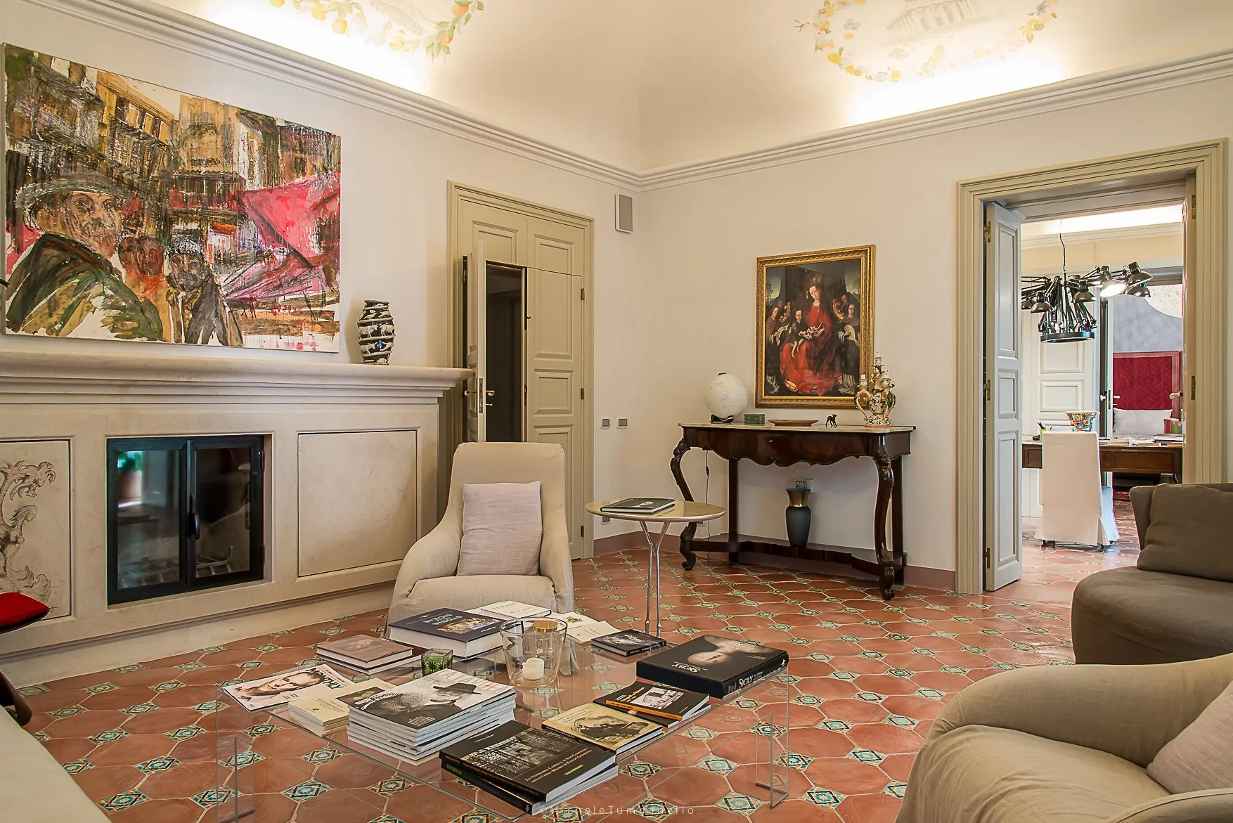 Living room with beige walls, tiled floor, cream-colored armchair with cushion, glass coffee table with magazines, and artwork on the walls. A doorway leads to a dining area with a chandelier, and another doorway to a hallway.