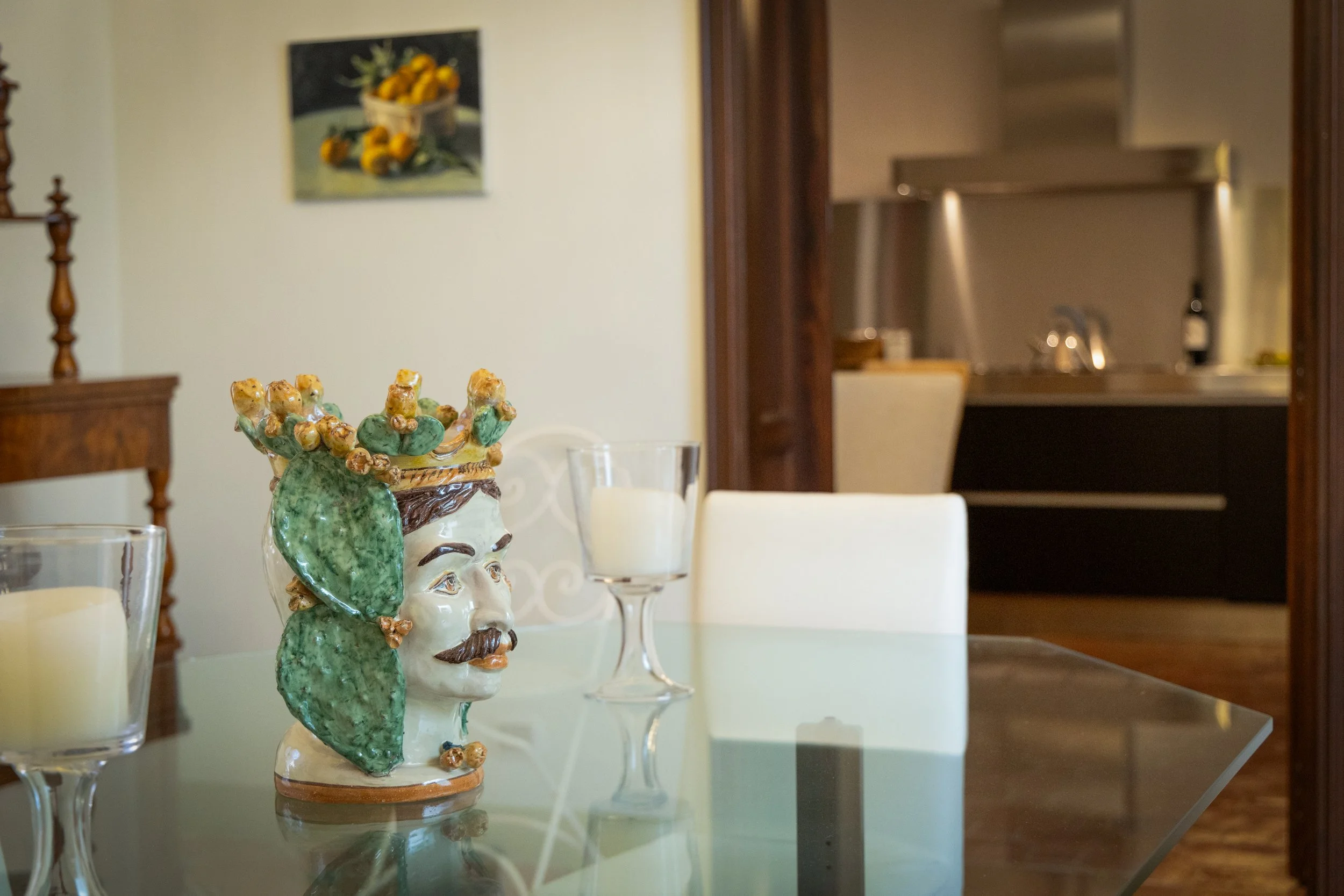 Decorative ceramic bust of a man with a mustache, wearing a crown of grapes and leaves, on a glass dining table with candles, in a cozy home kitchen and dining area.