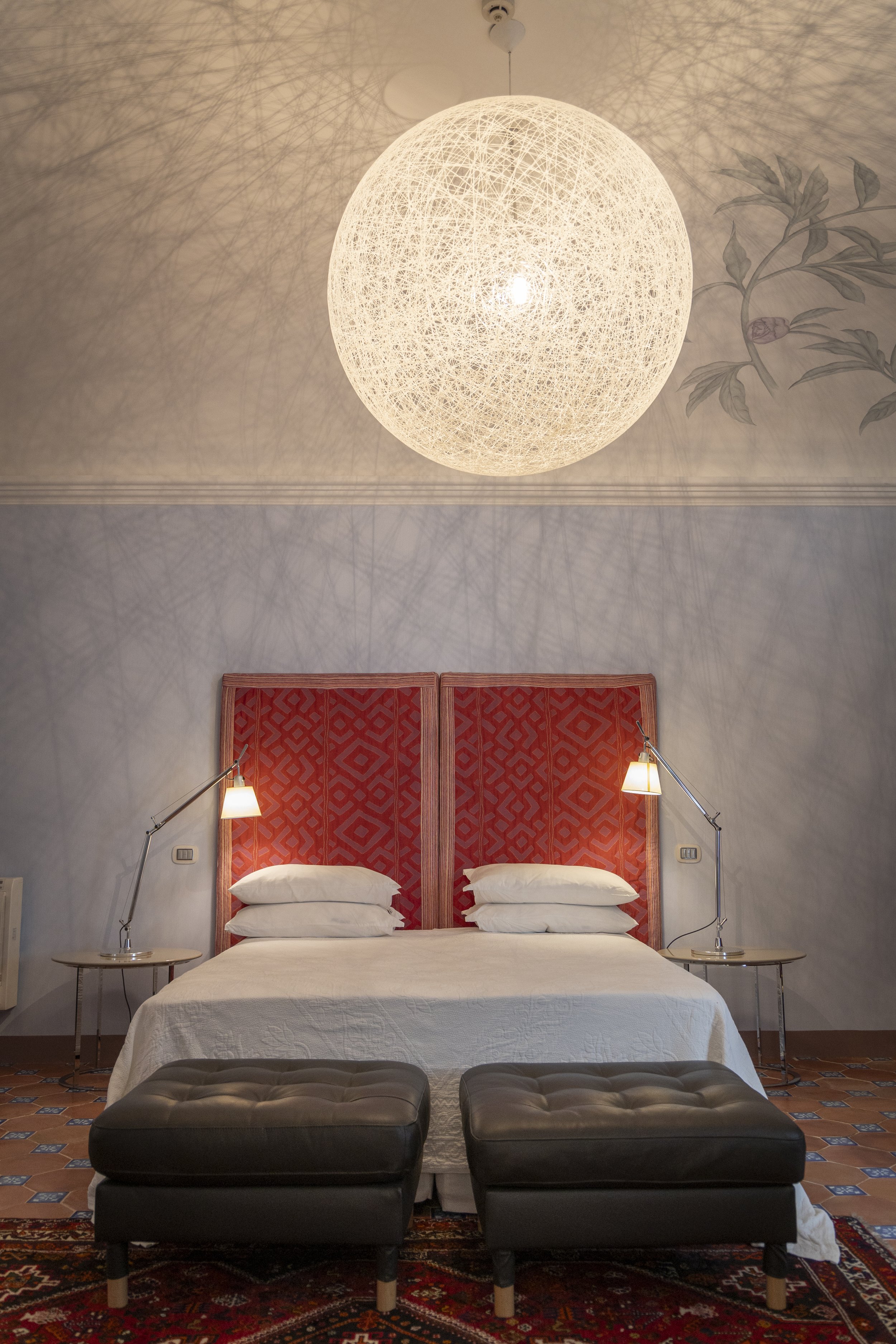 A bedroom with a large spherical ceiling light casting shadows on the wall, a bed with a white quilt and stacked pillows, a red upholstered headboard, two small side tables with adjustable lamps, a brown leather ottoman at the foot of the bed, and pa