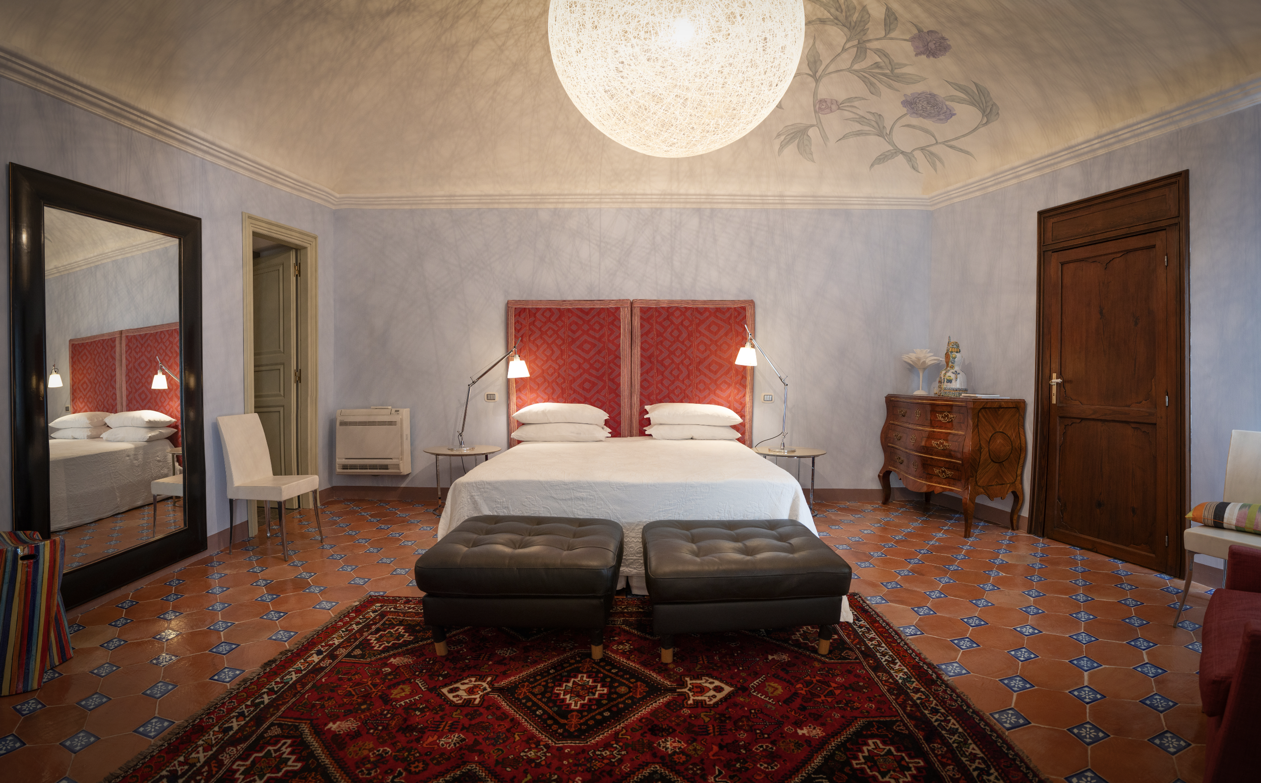 A hotel room with a double bed that has a red patterned headboard, two bedside lamps, and white bedding. There are two black ottomans at the foot of the bed, a large mirror on the left, a wooden dresser on the right with decorative items, and a rug on a terracotta-tiled floor. The walls are light blue with floral accents on the ceiling.