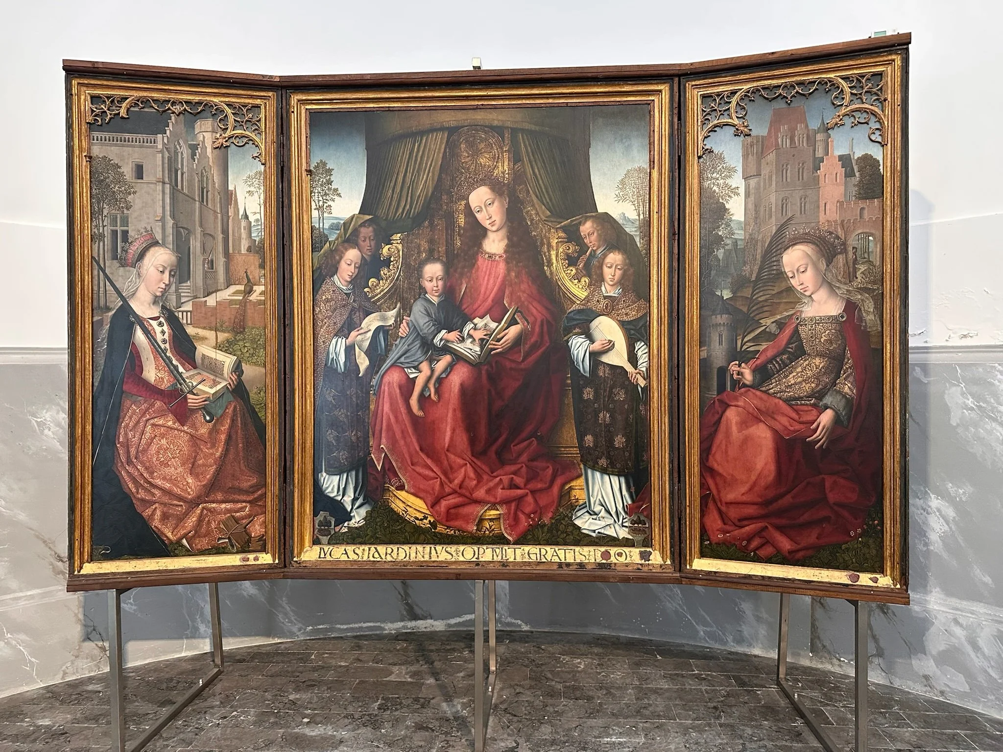 A Renaissance triptych painting depicting the Madonna and Child in the center, with four saints or apostles surrounding them. The left panel shows a female saint holding a book and a sword, while the right panel shows a seated female saint holding a palm branch. The background features architectural elements and a landscape.