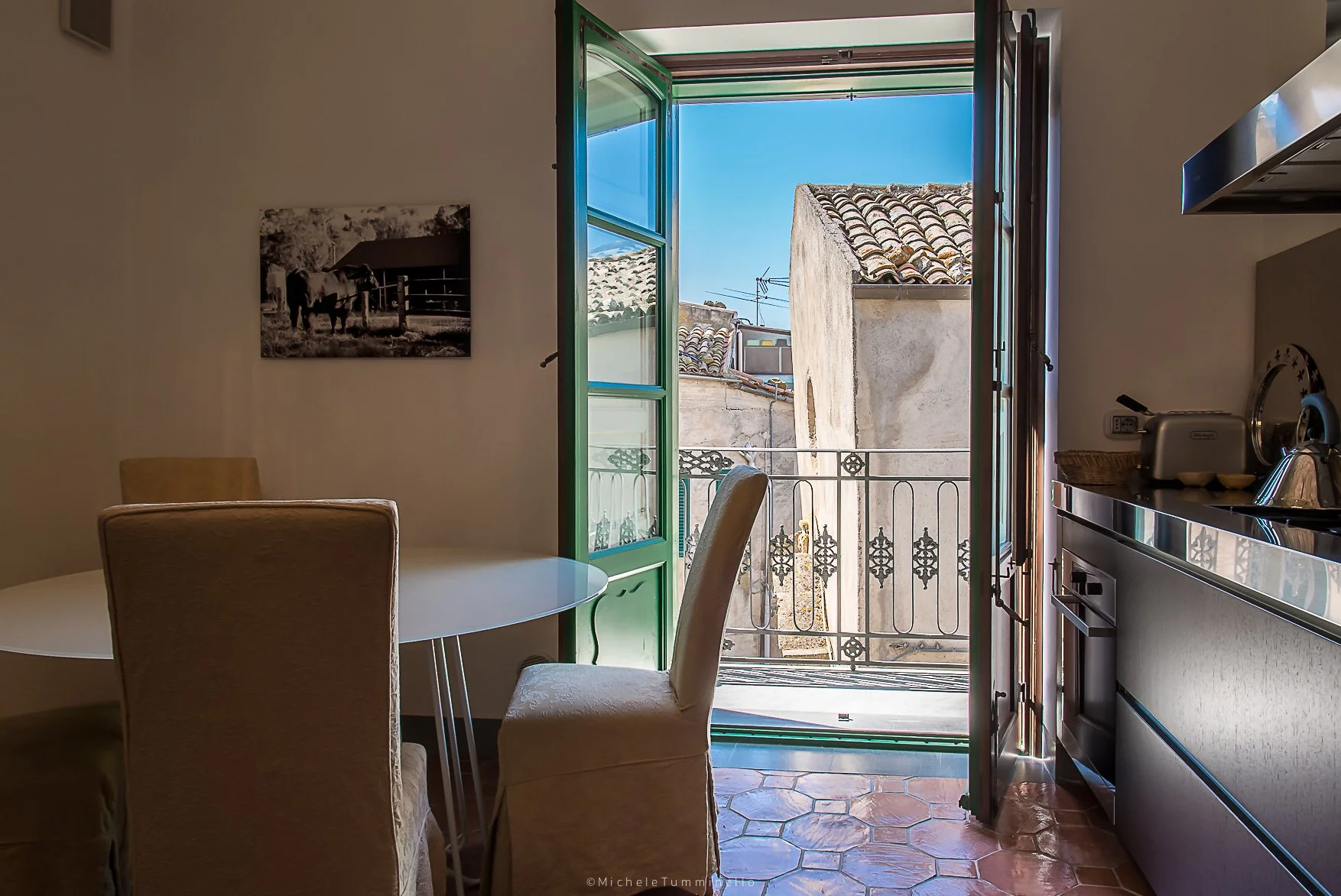 Kitchen with open balcony door showing rooftops and blue sky, dining table with chairs, small kitchen appliances, and terracotta tile flooring.