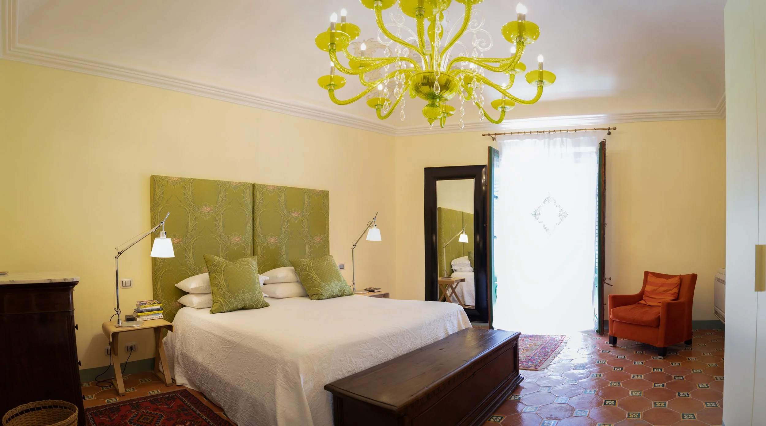 A bedroom with a large bed with green padded headboard, white bedding, and multiple green pillows. Beside the bed is a nightstand with a lamp and books. There is a mirror with a black frame near the window, which has sheer white curtains. An orange a