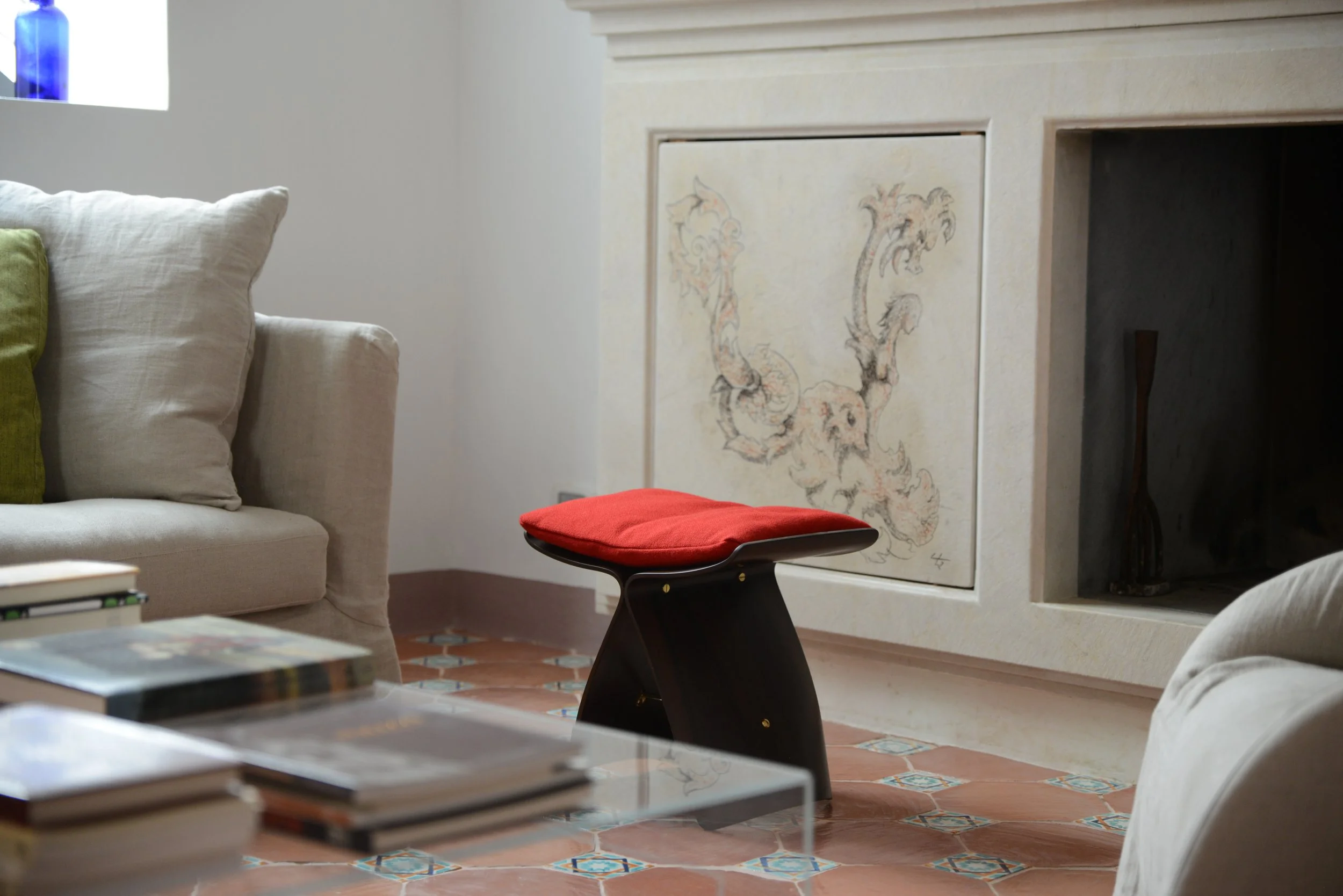 Living room with a beige couch, white and green pillows, a glass coffee table with books, a red cushioned stool, a fireplace with a decorative panel, and terracotta tiled floor.