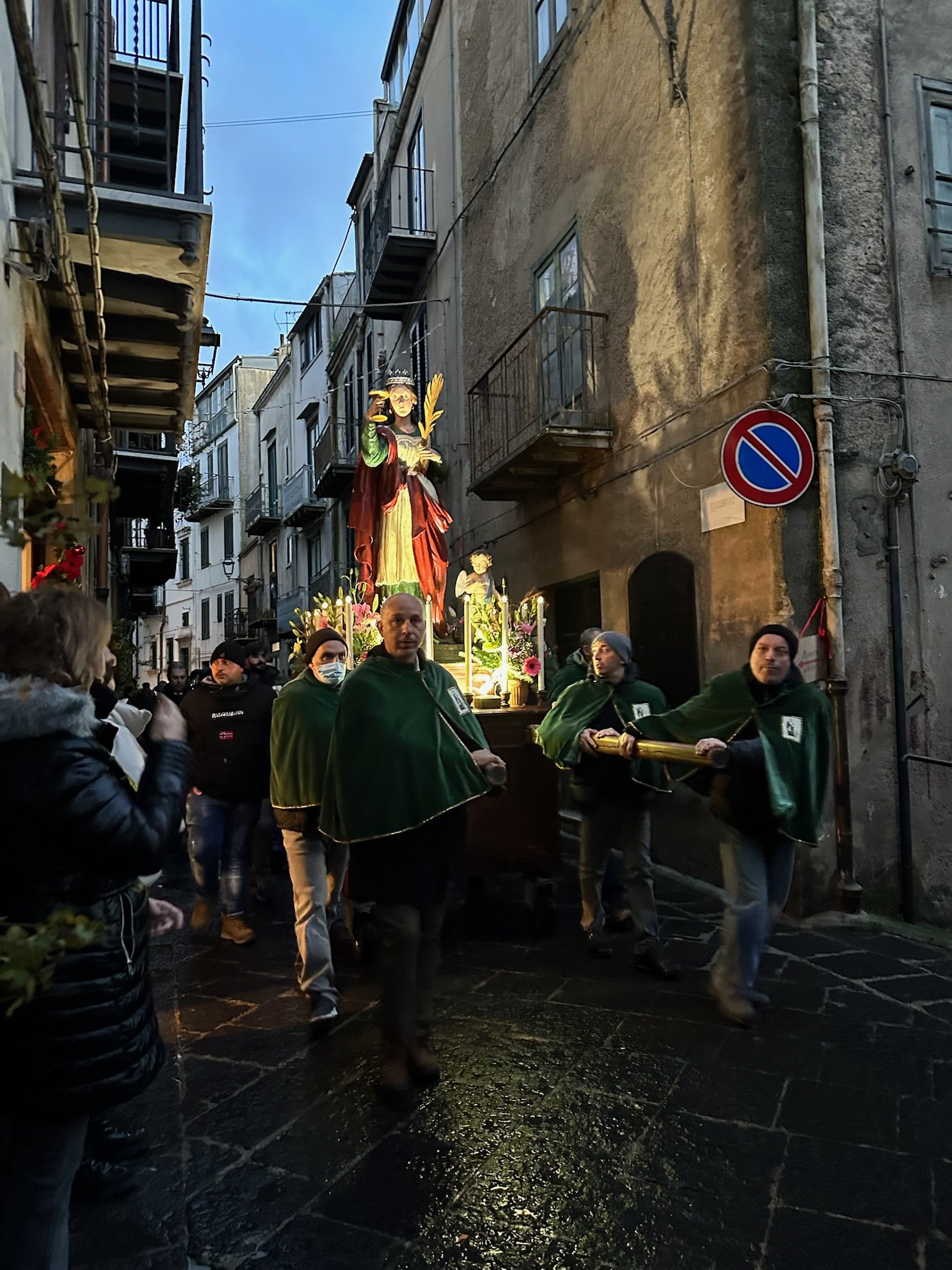 People participating in a religious procession, carrying a decorated float with a statue of a saint on a narrow street, some wearing capes and face masks, ambient evening light.