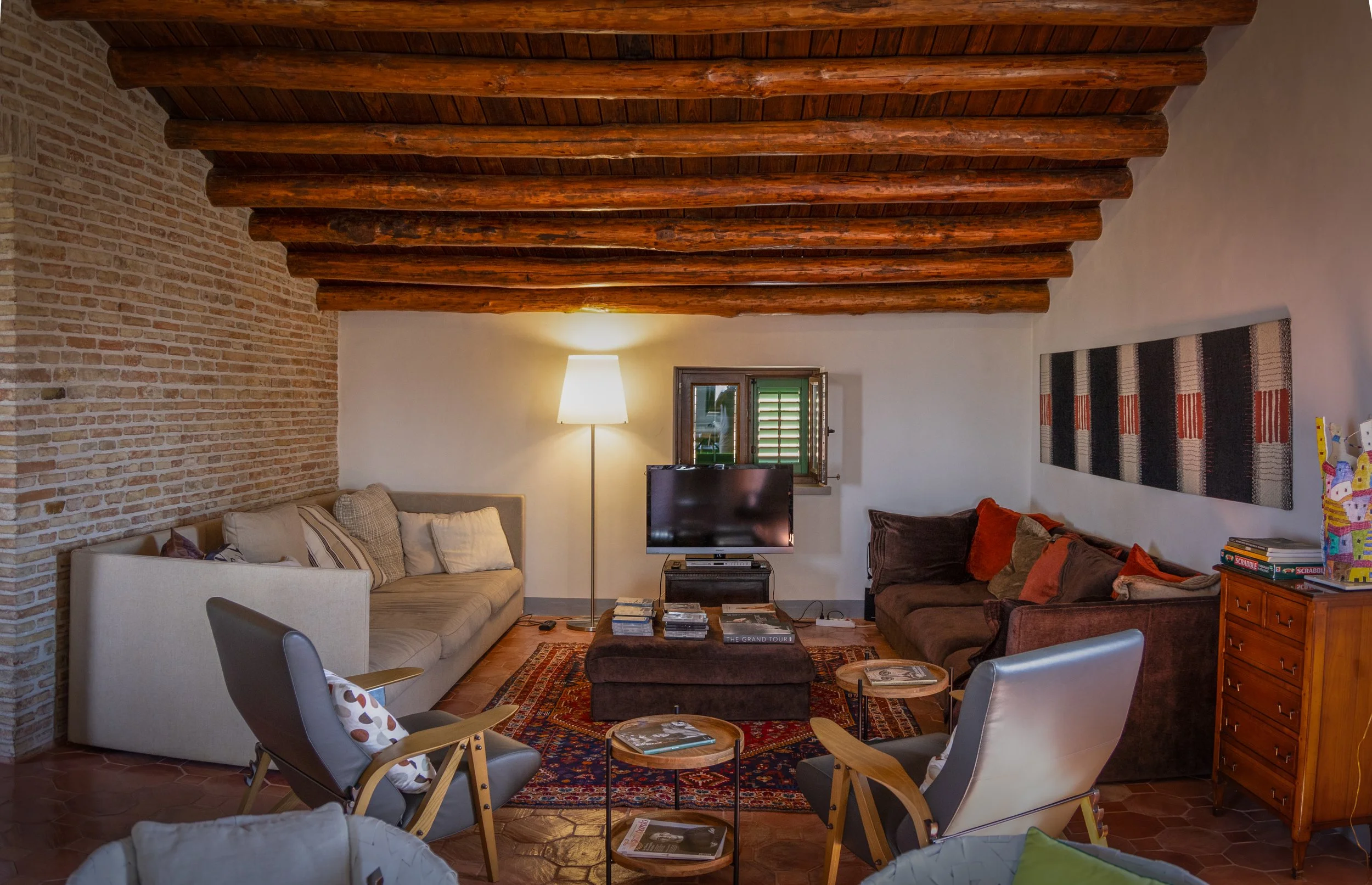 Cozy living room with exposed wooden beam ceiling, brick wall, and cushioned seating with throw pillows. Includes a small window, a TV, a floor lamp, and a wooden sideboard with books and decorative items.