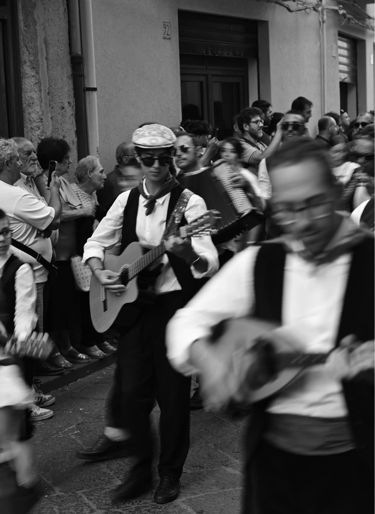 Black and white photo of street musicians playing guitars during a crowded outdoor event, with onlookers lining the sidewalk.
