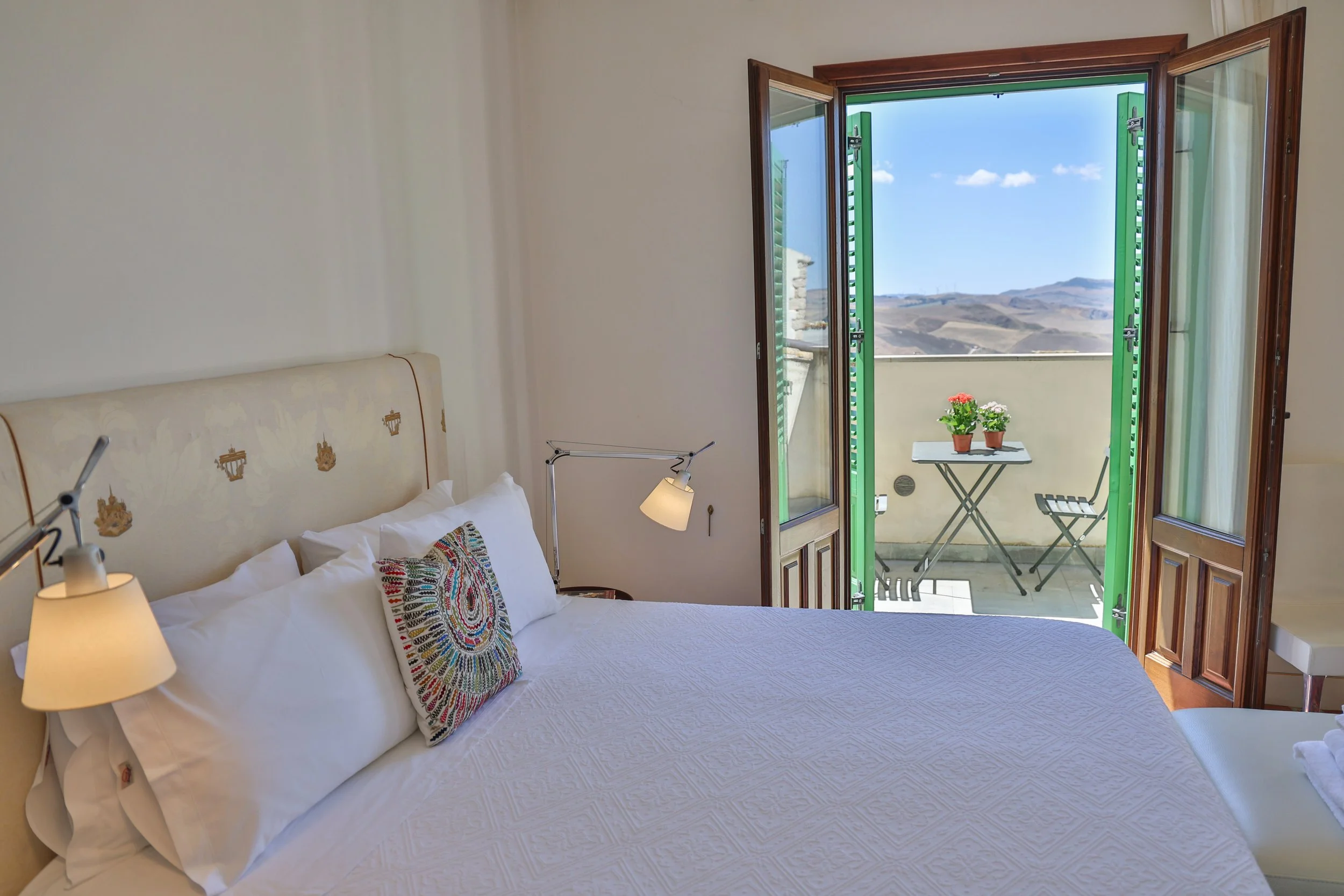 A bedroom with a bed, white bedding, and a colorful pillow. A door opens to a small balcony with a table and two potted plants, overlooking a landscape of rolling hills under a blue sky.