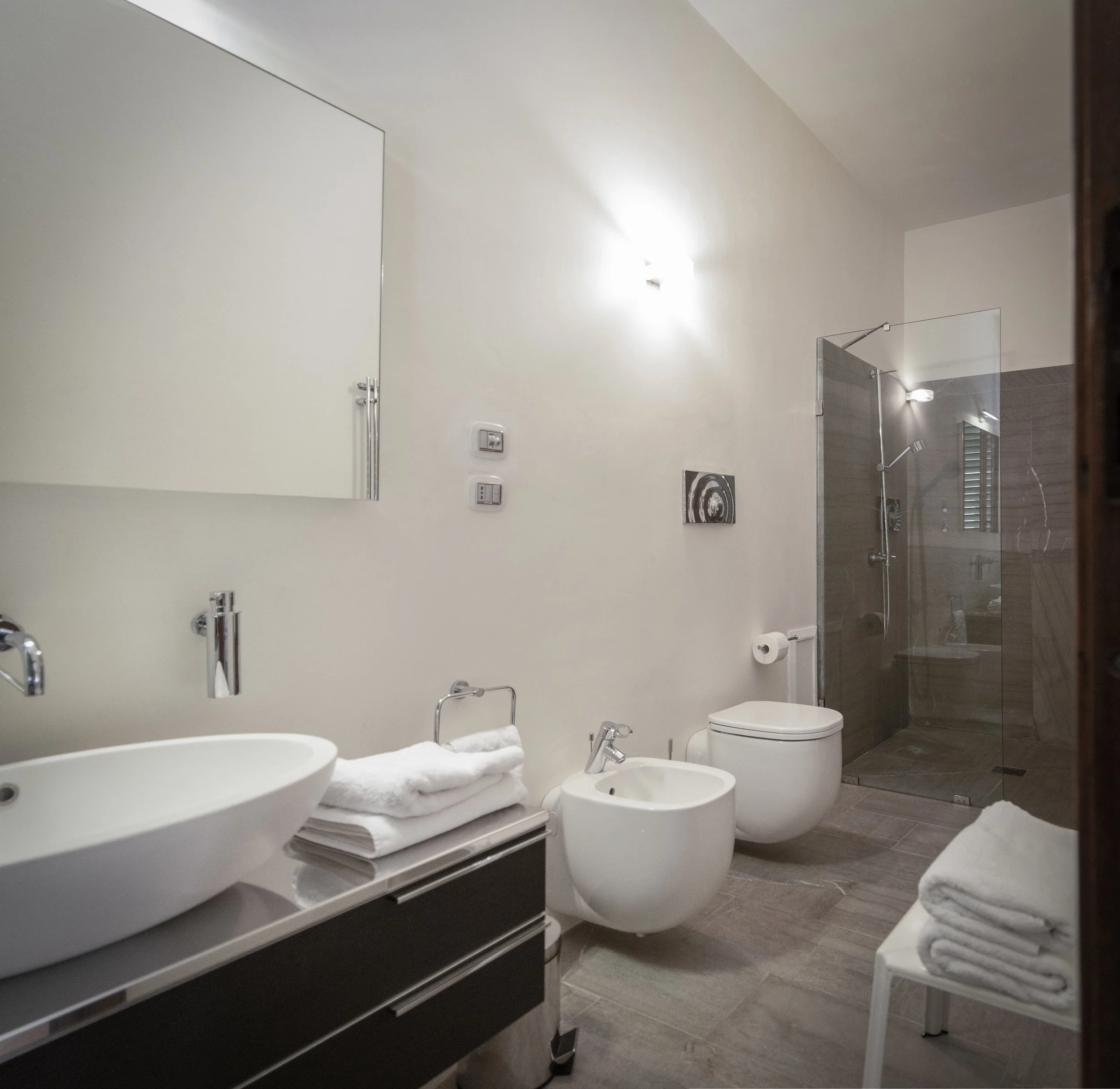 Modern bathroom with a white vanity, vessel sink, bidet, toilet, and walk-in shower with glass door and gray tiles.