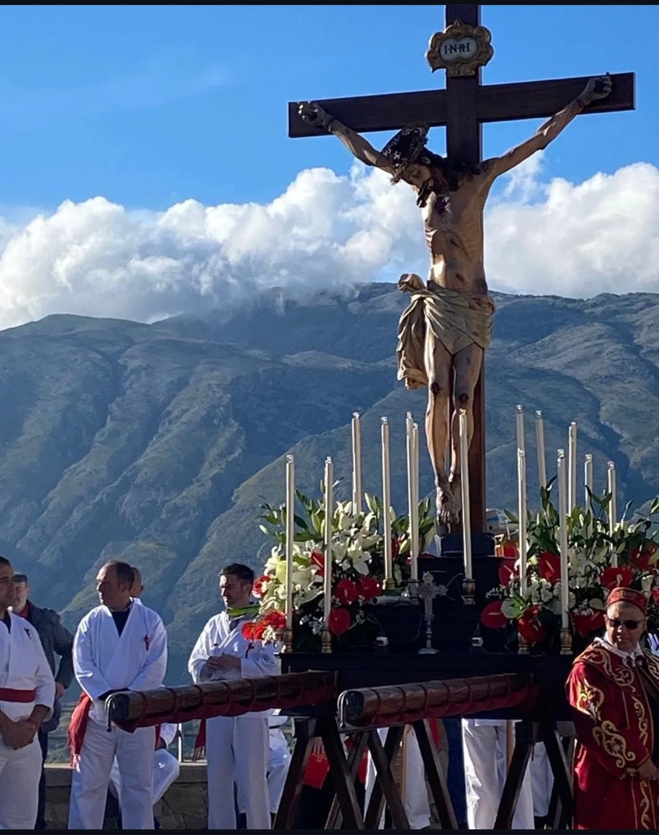 A religious procession with a large crucifix depicting Jesus Christ, surrounded by flowers and candles, on a platform carried by people in traditional ceremonial attire, set against a backdrop of mountains and clouds.