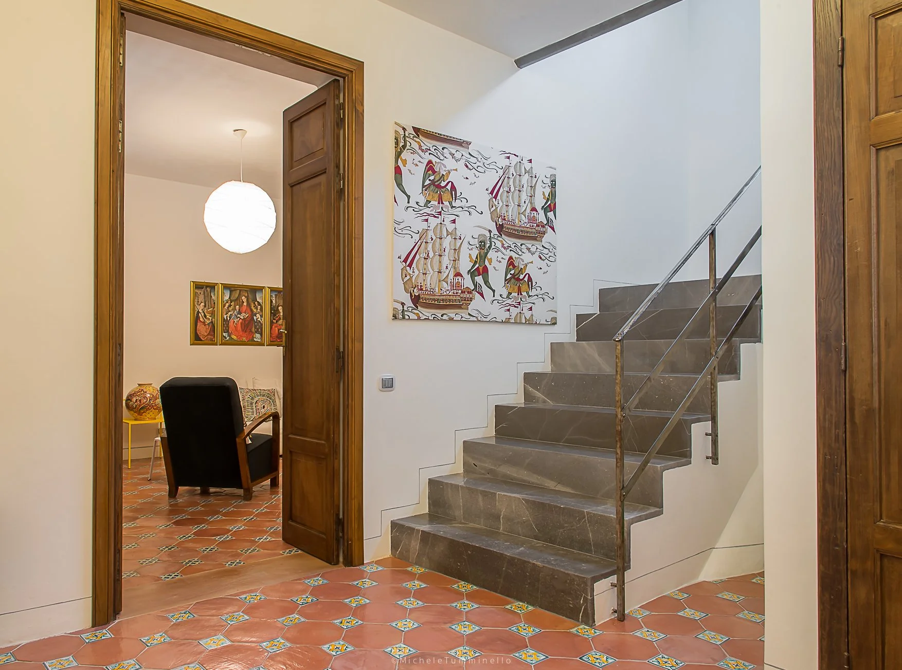 View of a staircase with dark stone steps and metal railing in a hallway, next to white walls with artwork, leading to an open room with terracotta tiled floor, a black armchair, colorful painting, and a table with a decorative vase.