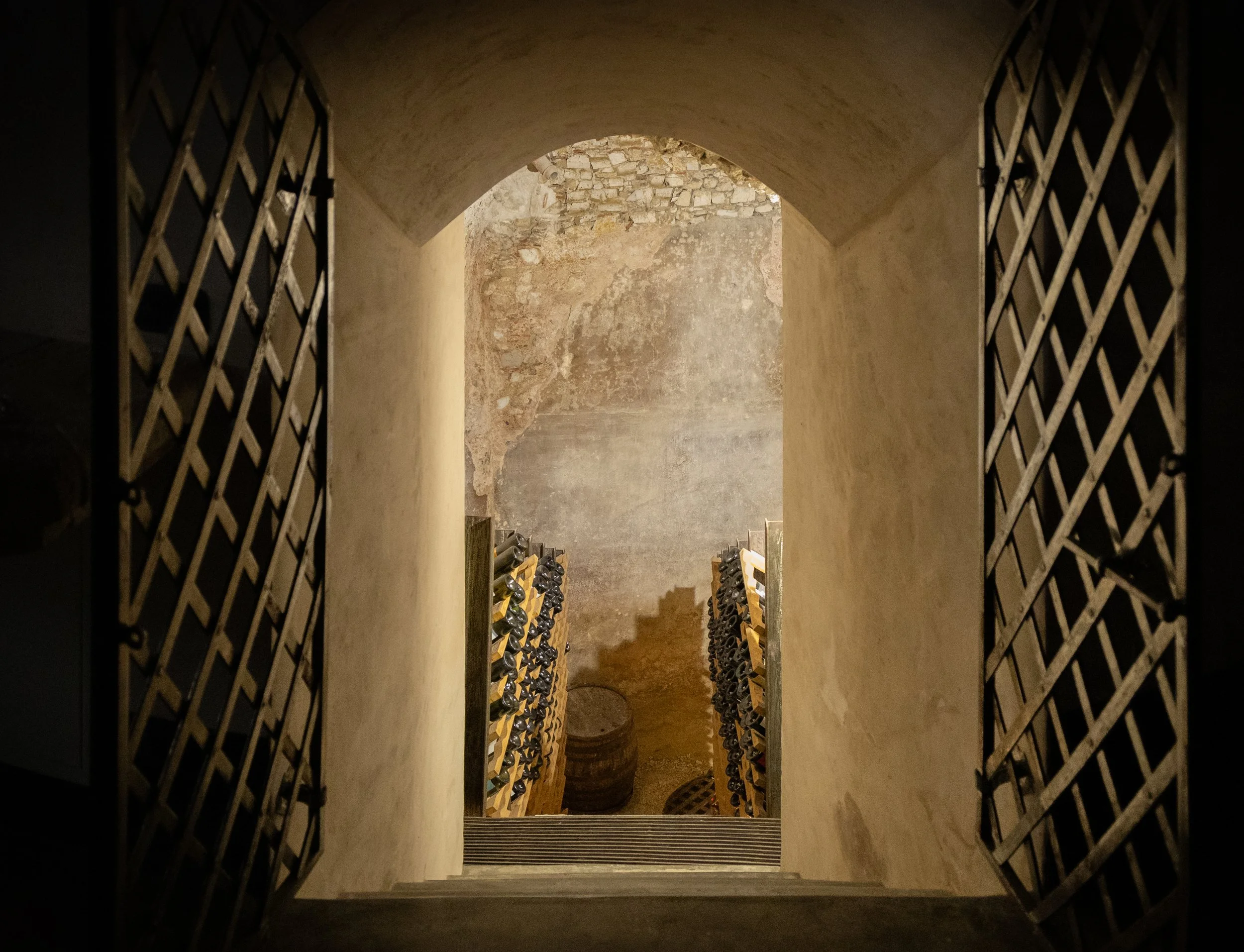 View through an arched doorway into a wine cellar with wine racks on each side filled with bottles.