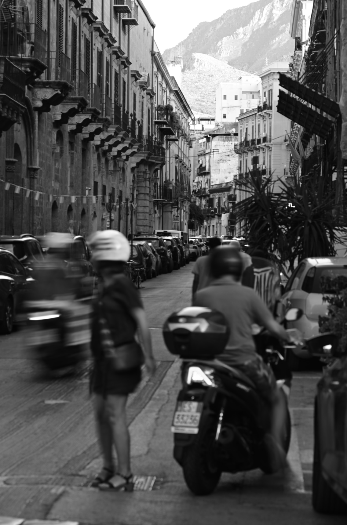 A black and white street scene with cars parked along the curb, a motorcycle with a woman in a skirt standing nearby, and a man on a scooter, with buildings and mountains in the background.
