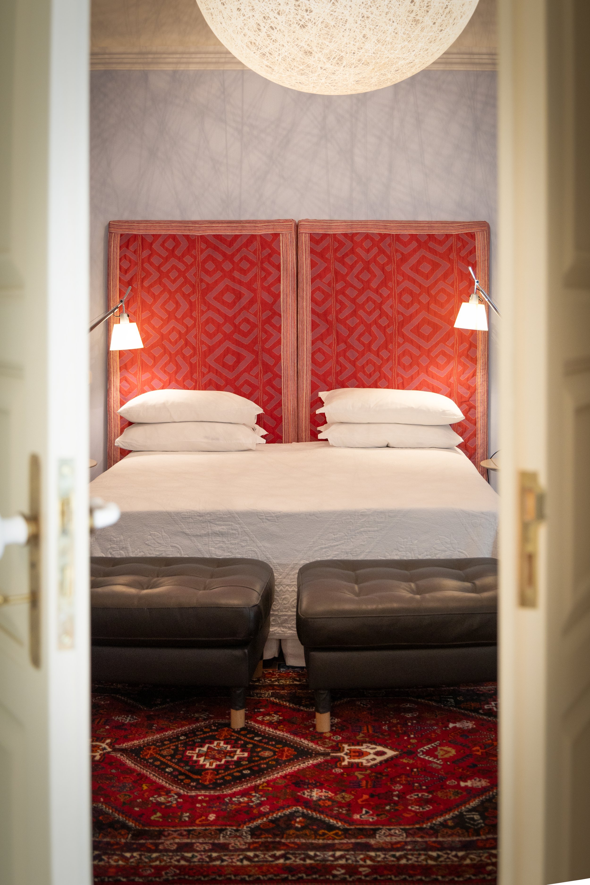 View of a hotel room with a bed that has a patterned headboard, four white pillows, and two wall-mounted reading lamps. Two black upholstered ottomans sit at the foot of the bed, and a red patterned rug is visible on the floor. The photo is taken fro