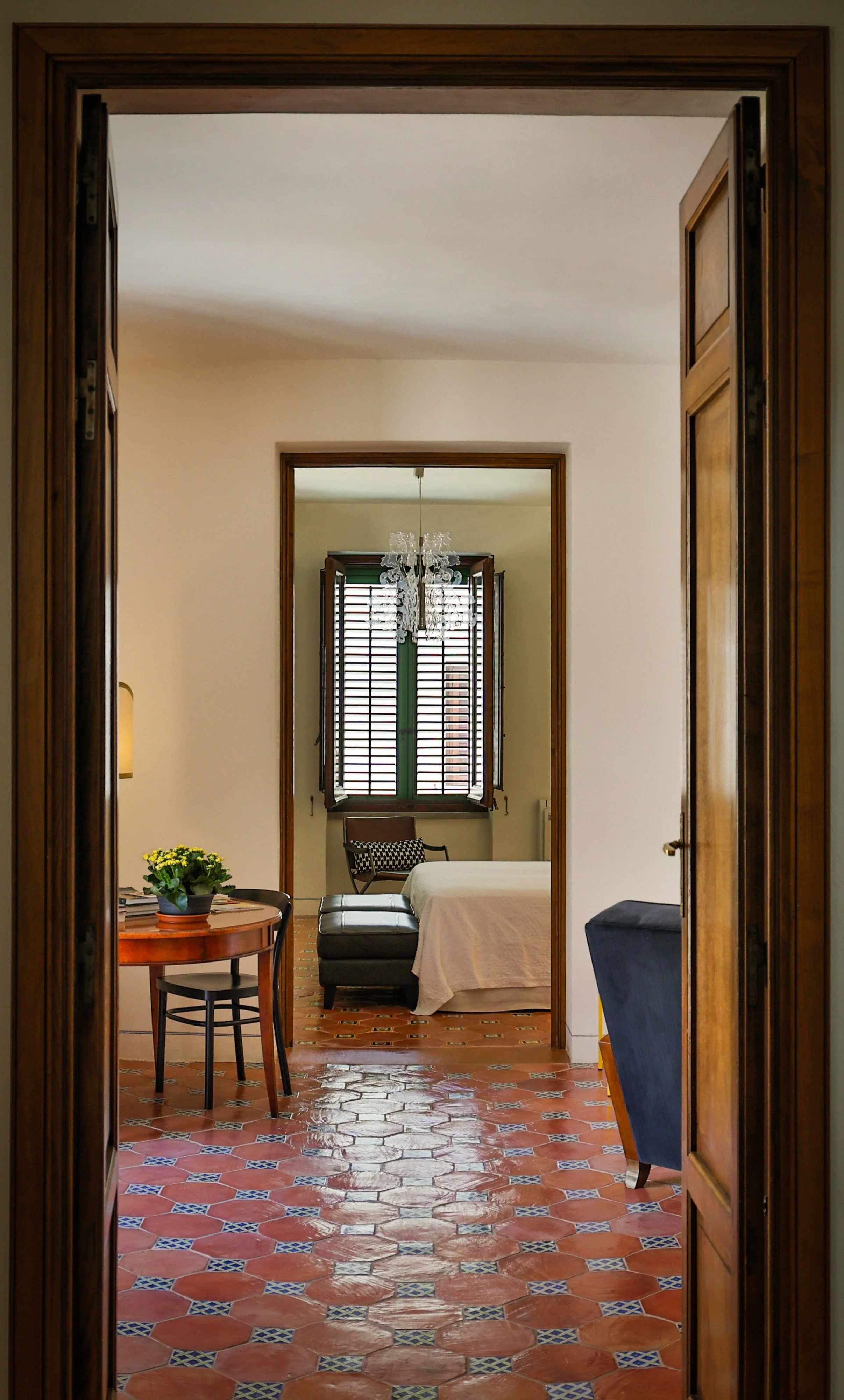 View through doorway into a bedroom featuring terracotta tiled flooring, a bed with white bedding, a window with green trim and wooden shutters, a chandelier, a black leather bench at the foot of the bed, a small round side table with a potted plant,