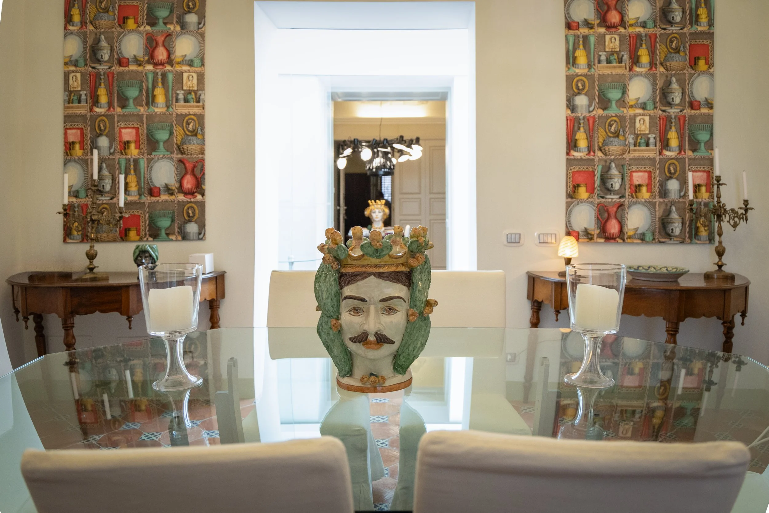 A decorative ceramic head sculpture with a crown on top as a centerpiece on a dining table. The dining room has traditional wooden sideboards, colorful wall hangings, and a doorway with white curtains in the background.