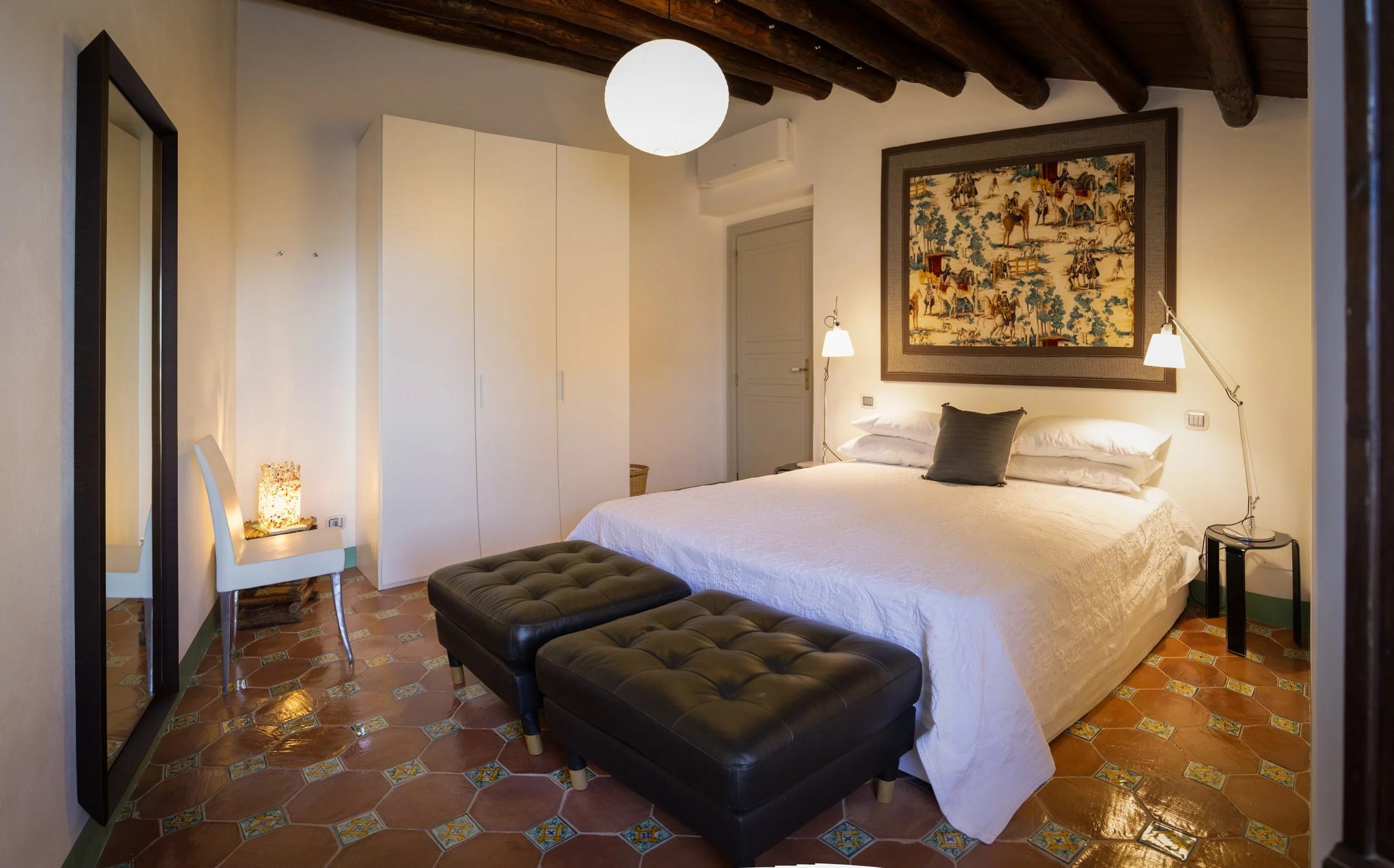 Cozy bedroom with a large white bed, brown tufted ottomans at the foot, a framed tapestry above the bed, a wardrobe, a white chair with a lamp, and a ceiling with exposed wooden beams.