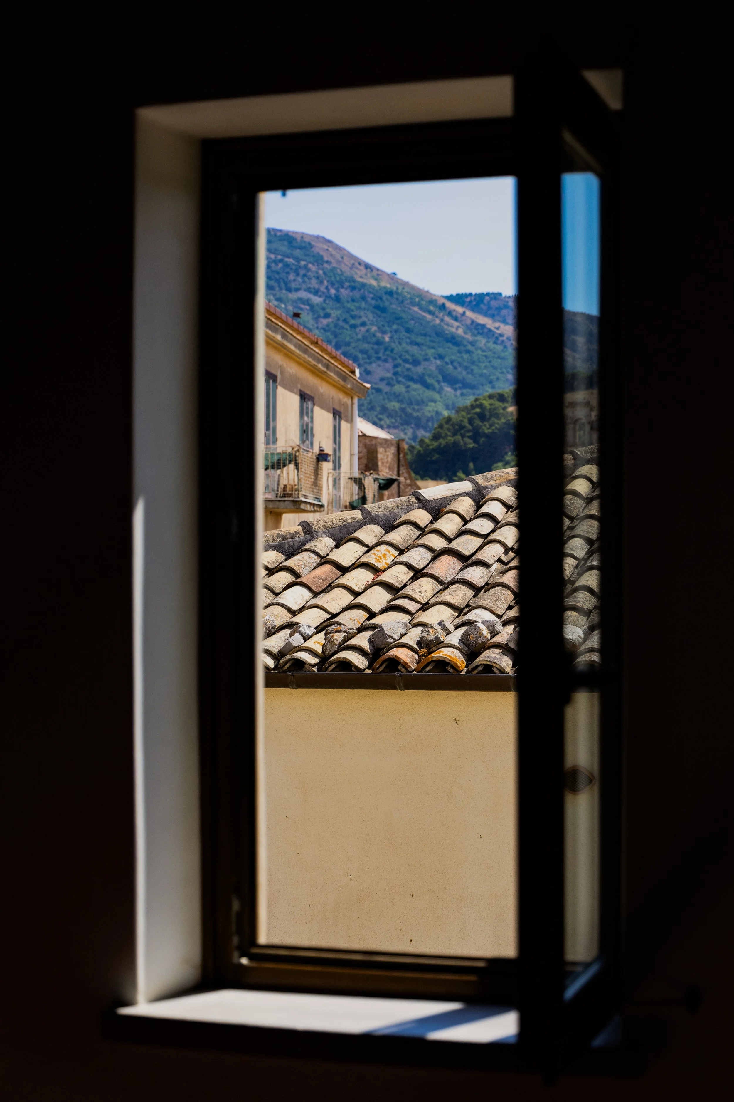 View through an open window showing a tiled roof, a building, and a mountain in the background.