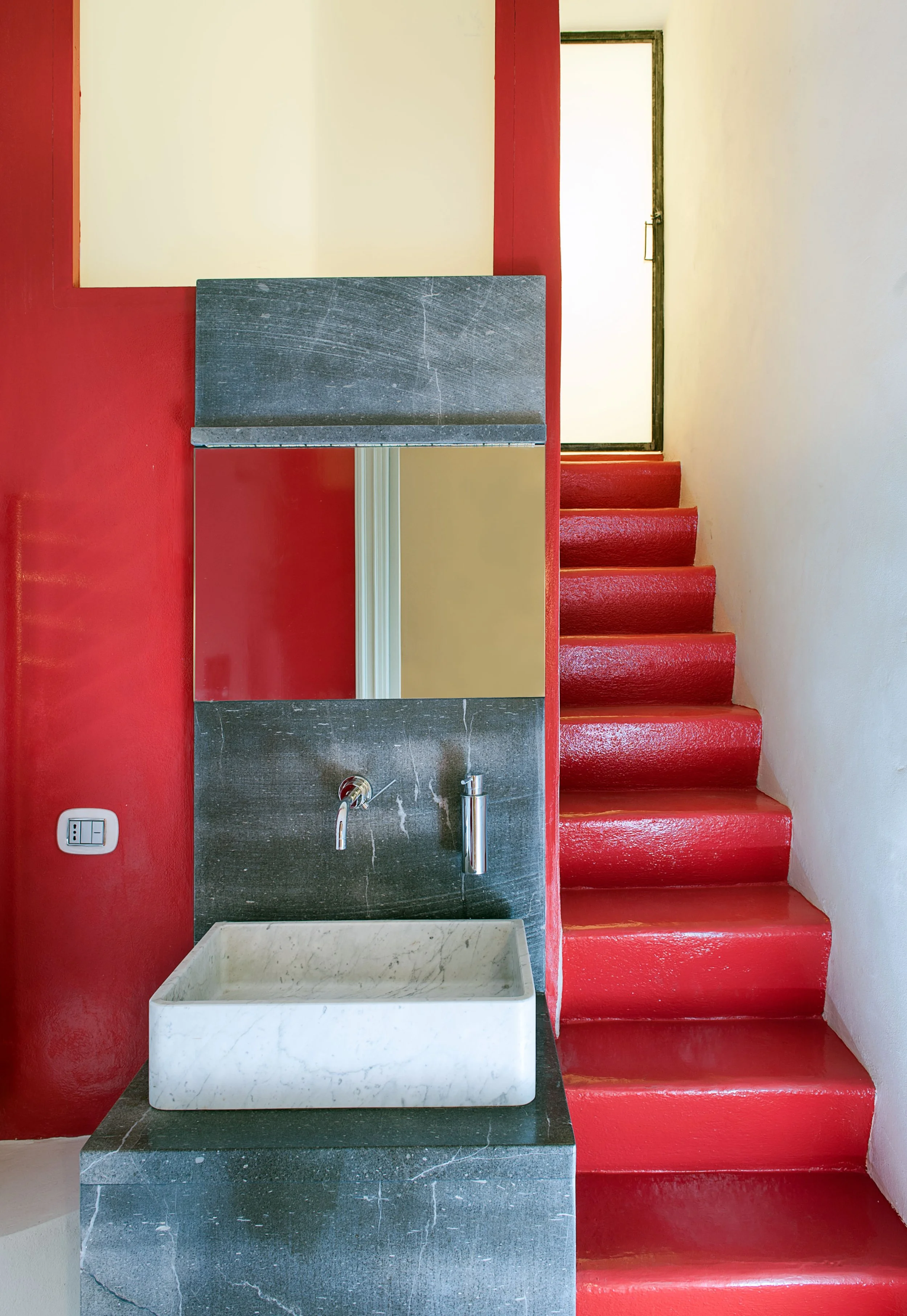 Interior view of a small space with a red staircase on the right, a dark gray sink with a white marble basin, a mirror, and a door with a frosted glass window at the top of the stairs.
