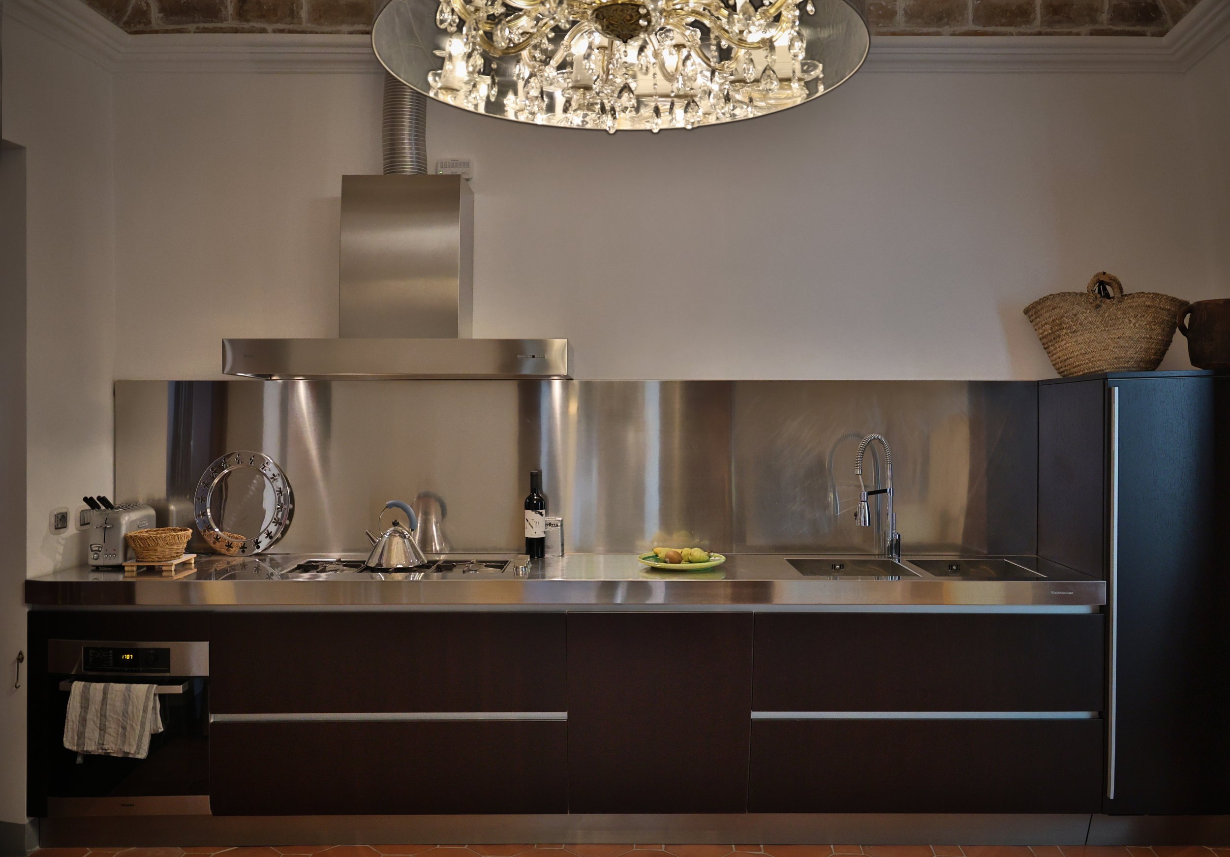 Modern kitchen with stainless steel countertops and dark cabinets, includes stove, microwave, sink, and decorative items like baskets and a plate of fruit.