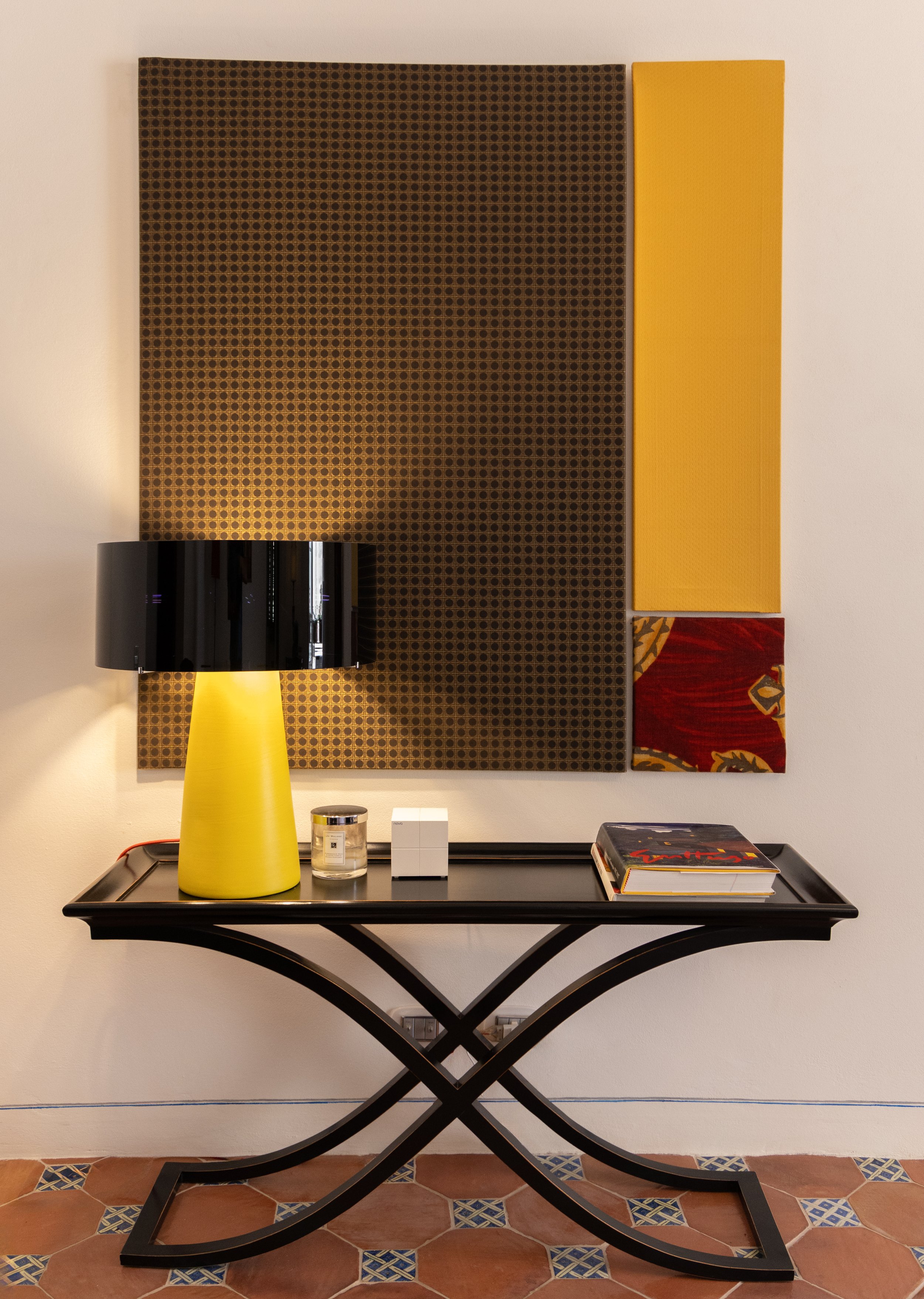 Decorative wall art with geometric patterns hangs above a black console table holding a yellow lamp, candle, small boxes, and a large book, in a well-lit room.