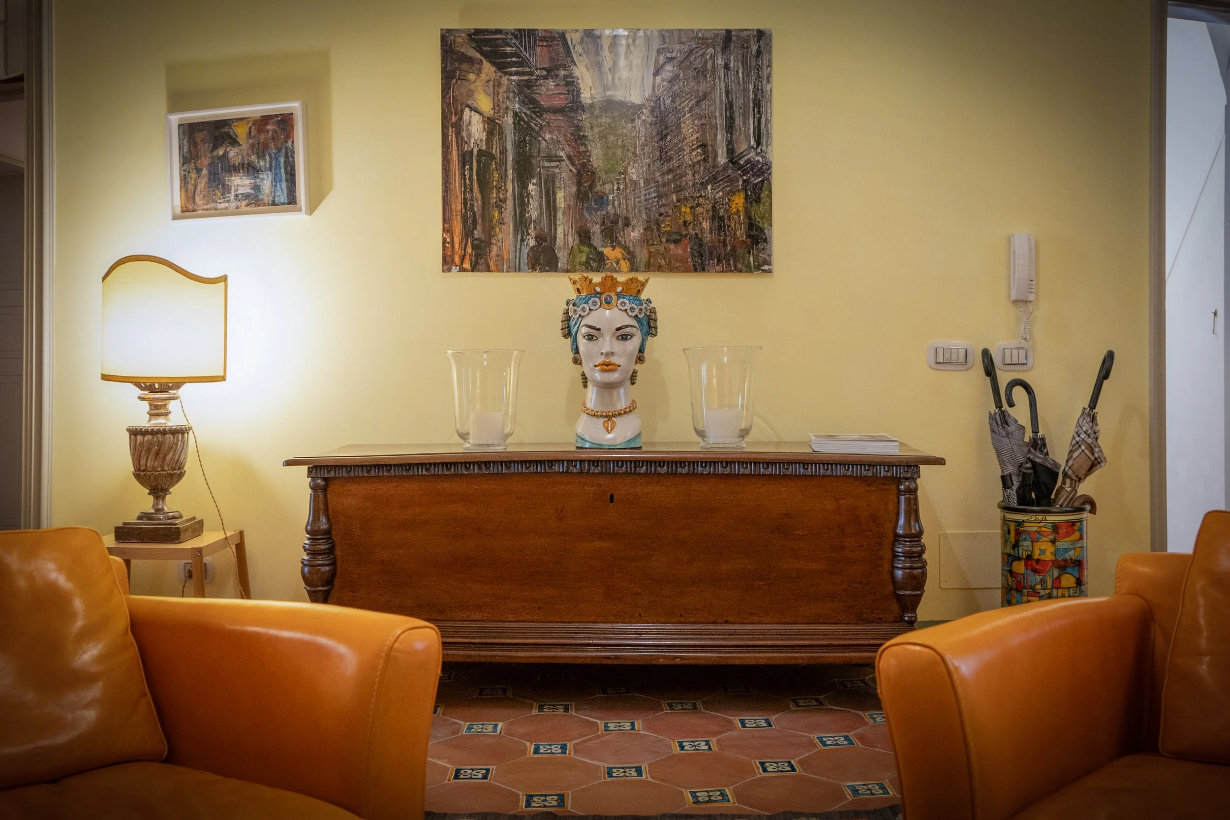 Living room with a wooden sideboard, a decorative bust, and two large candle holders on top, surrounded by leather armchairs, a floor lamp, and artwork on the walls.