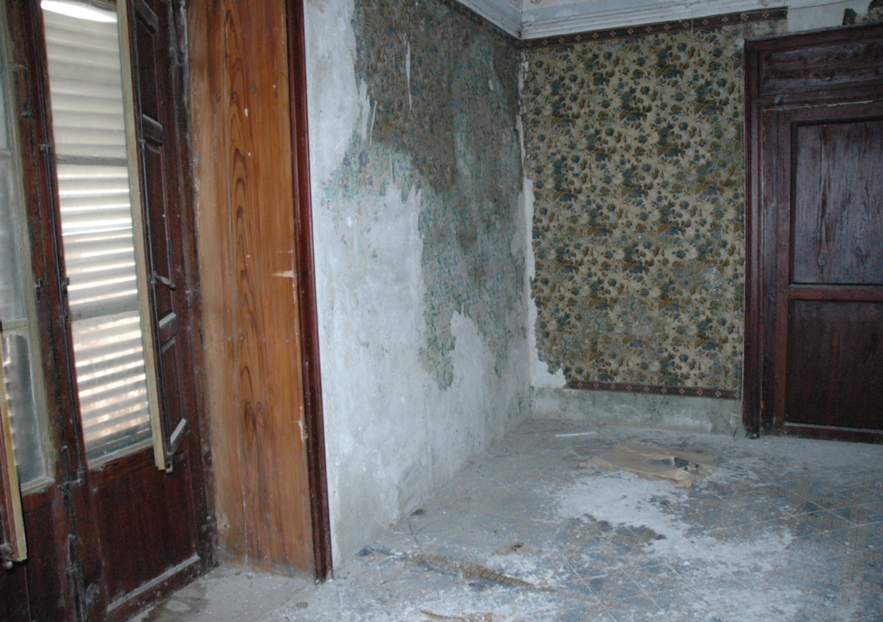 A room with damaged walls, peeling wallpaper, and a concrete floor with debris. There is a dark wooden door on the right and a window with closed shutters on the left.