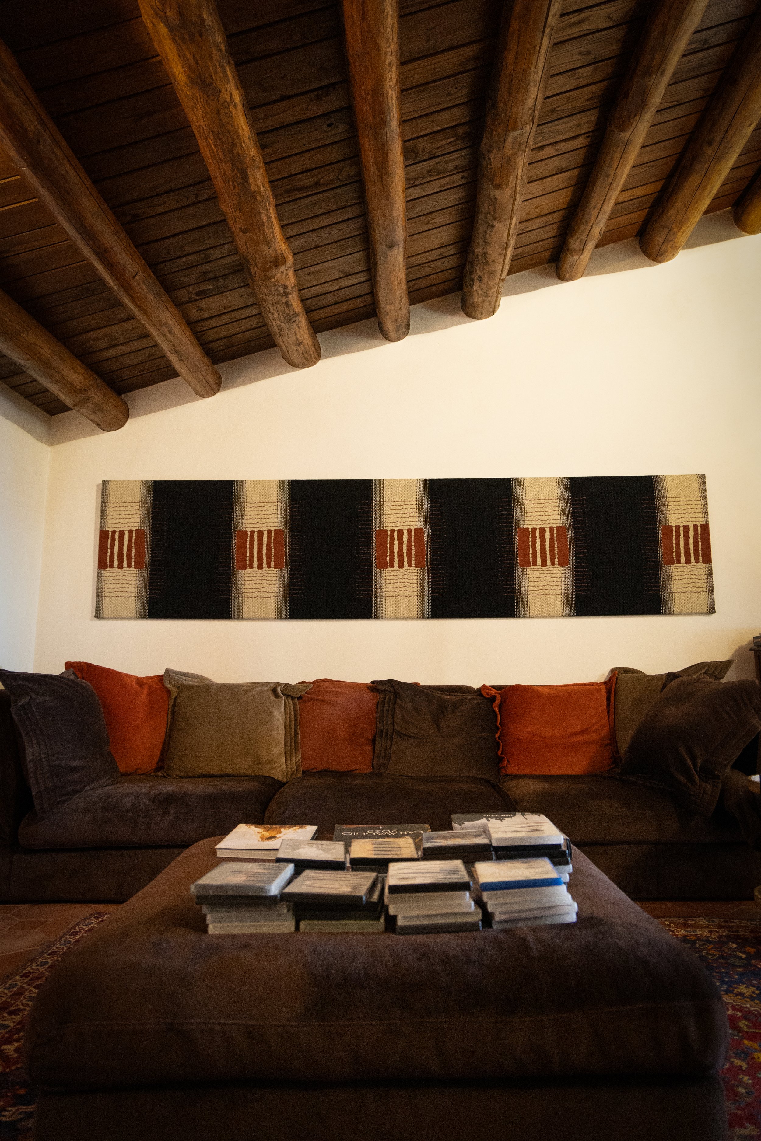 Living room with a brown sofa, colorful pillows, an ottoman with stacks of DVDs or books, rustic ceiling with exposed wooden beams, and a fabric wall hanging with checkered and striped patterns.