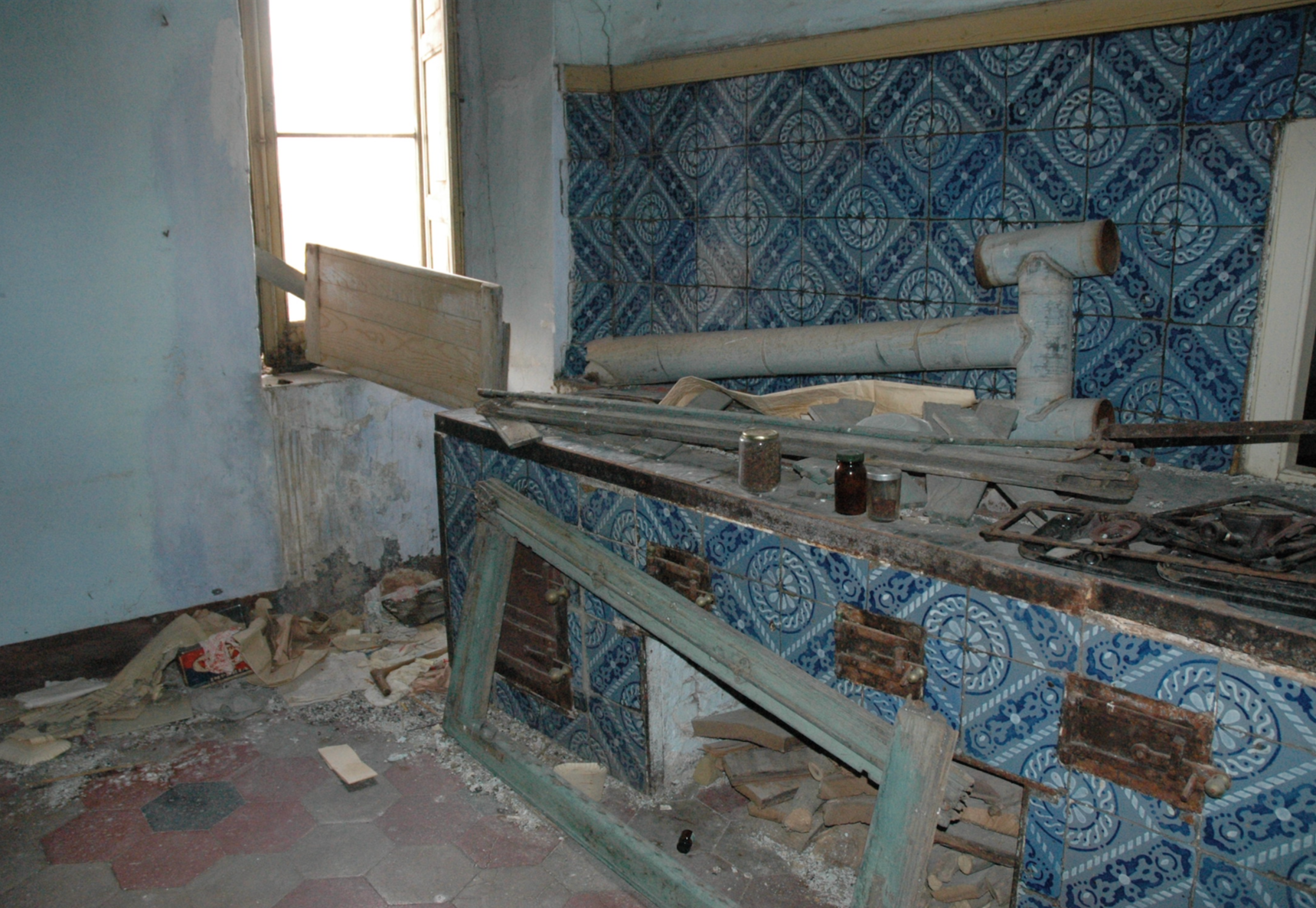 Abandoned and damaged kitchen with broken cabinets, debris on the floor, and a window with broken shutters, indicating neglect or vandalism.