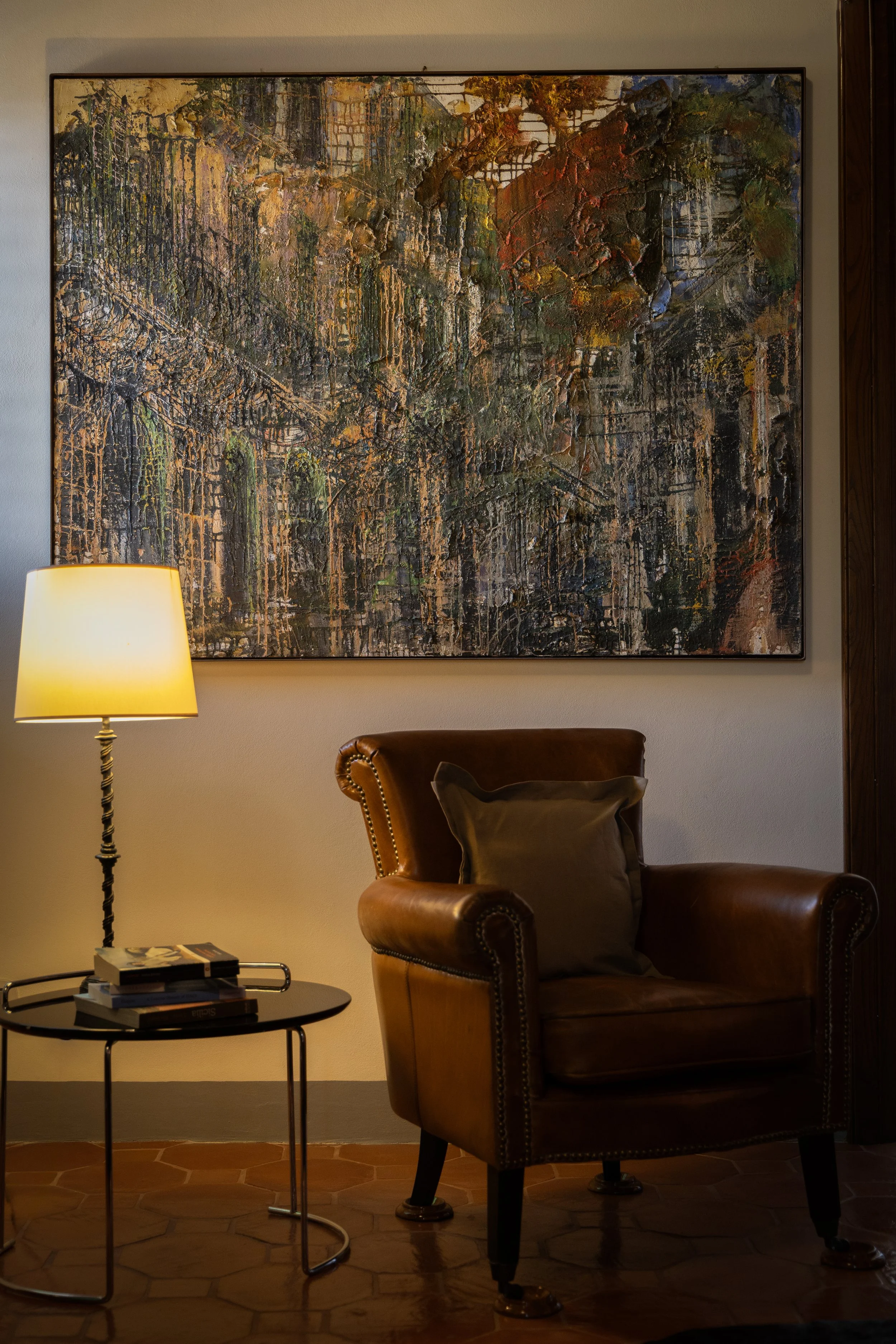 Cozy reading nook with brown leather armchair, dark side table with books, standing lamp, and abstract artwork on light-colored wall.