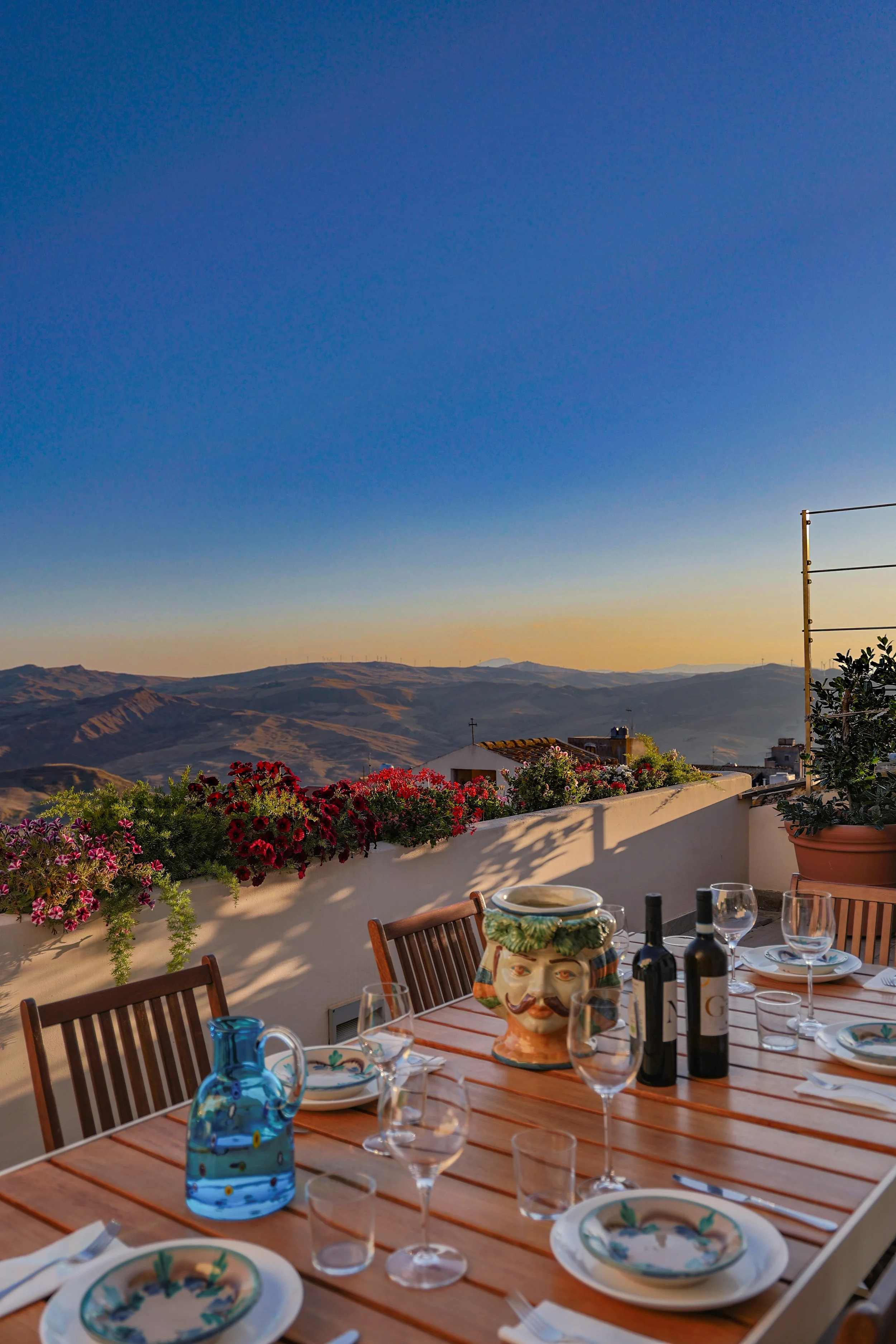 Outdoor dining table set on a terrace with hills in the background, decorated with flowers, wine bottles, glasses, and a colorful ceramic face planter.