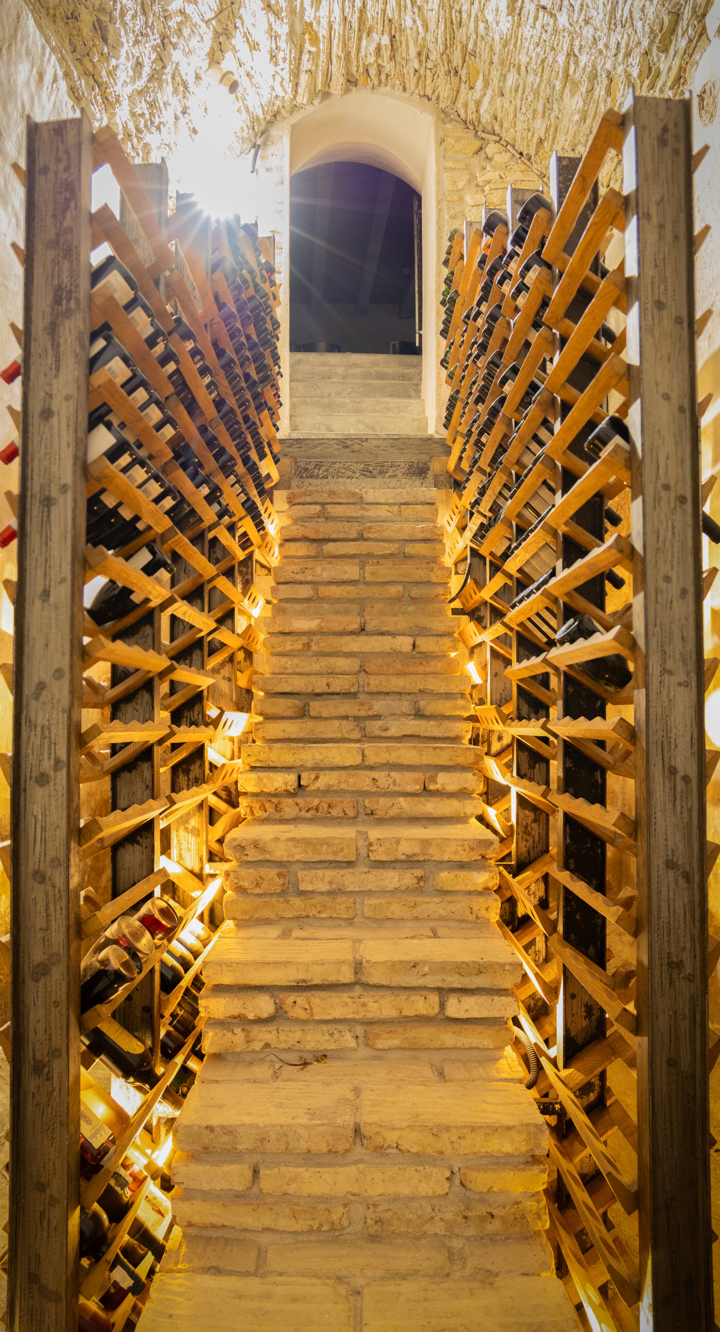 A wine cellar with brick stairs and wine bottles stored on wooden racks, illuminated by warm lighting.