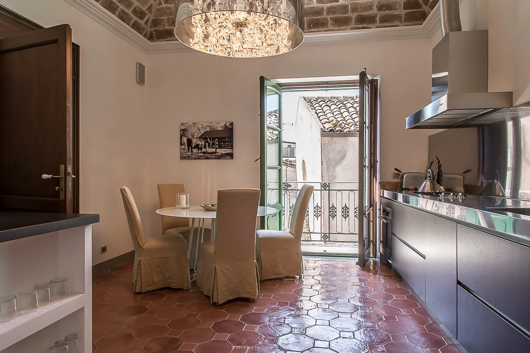 Kitchen with a round dining table and four beige upholstered chairs, an open doorway leading to a small balcony with black wrought iron railing, a wall-mounted photograph of a horse, and modern kitchen appliances, wooden cabinets, and tiled flooring.
