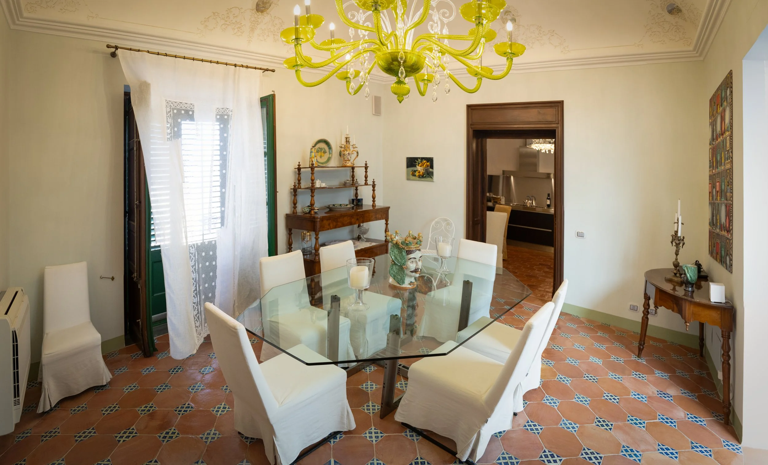 Dining room with a glass-top table surrounded by white chairs, a yellow chandelier hanging from the ceiling, and vintage furniture and artwork on the walls. Open door leads to kitchen.