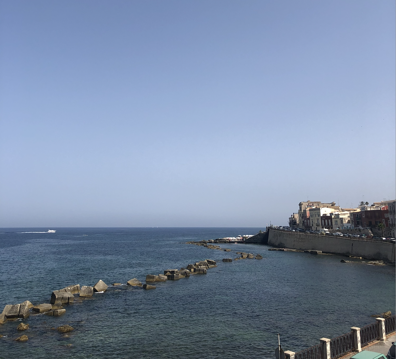 A coastal town with buildings along the shoreline, a clear blue sky, and calm ocean waters with a boat speedily passing in the distance.