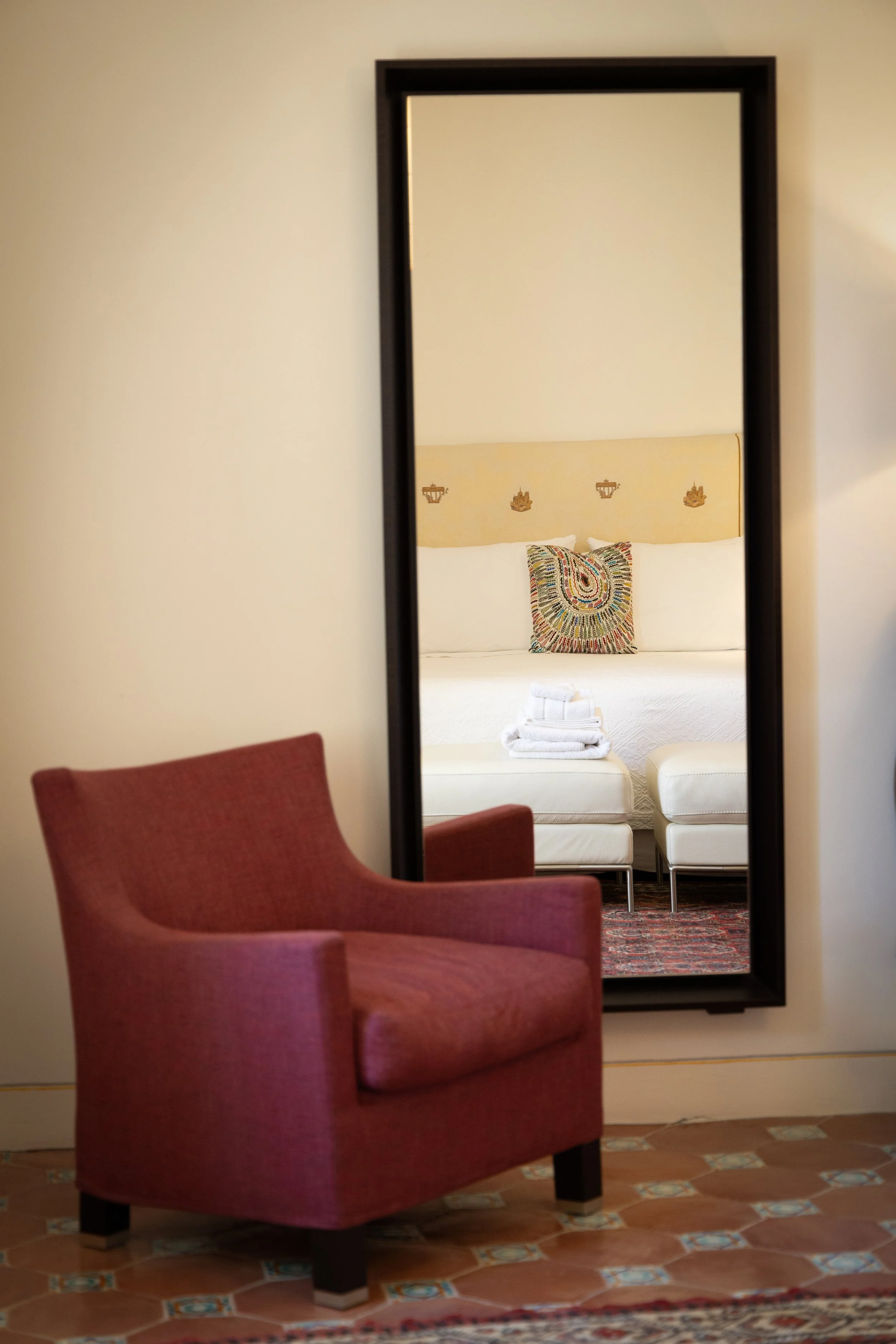 A room with a pink armchair in front of a large mirror reflecting a bed with white linens, decorative pillows, and folded towels.