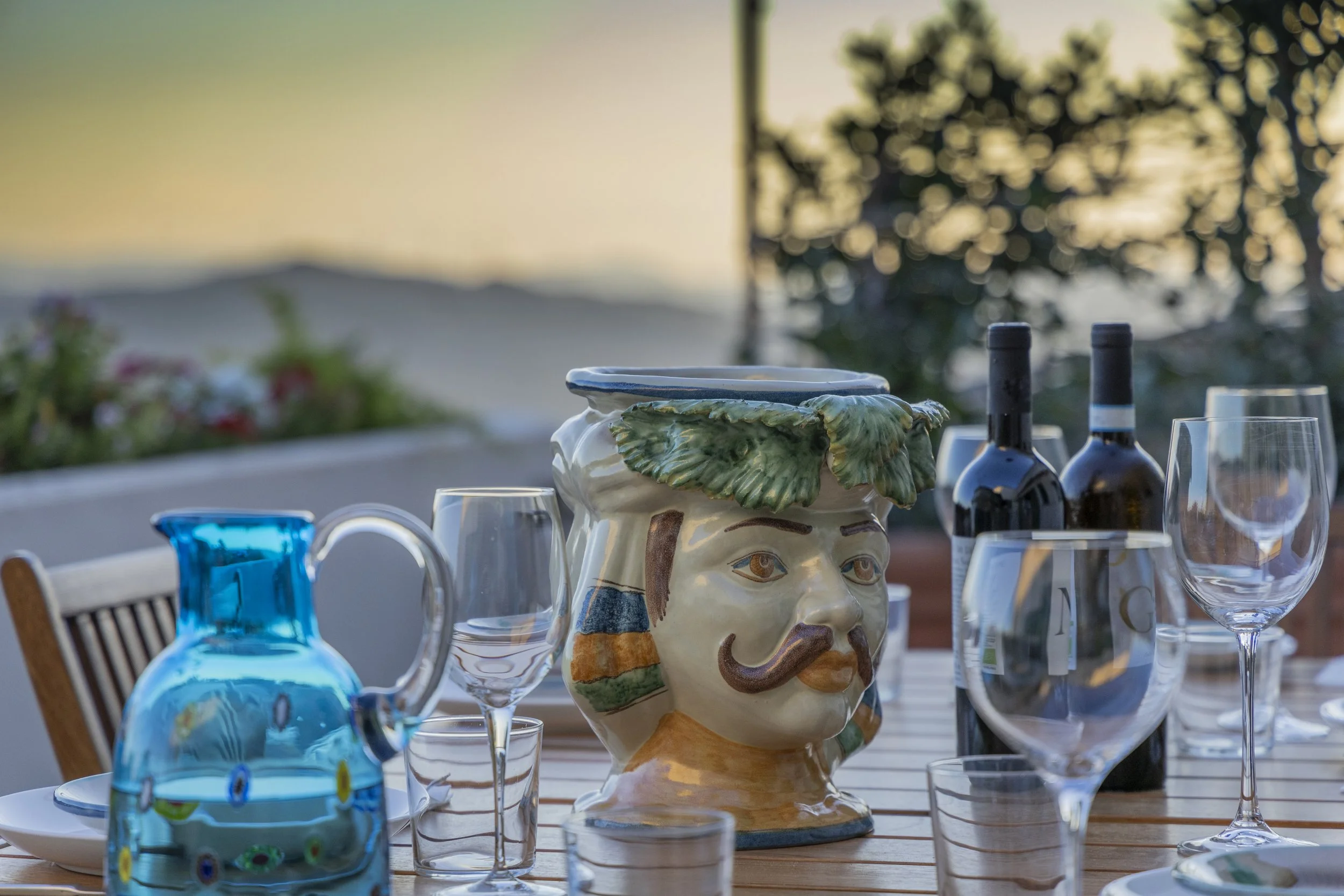 Outdoor dining table set with glasses, bottles of wine, and a decorative ceramic pitcher shaped like a man's face with a mustache and leafy green hair, during sunset.