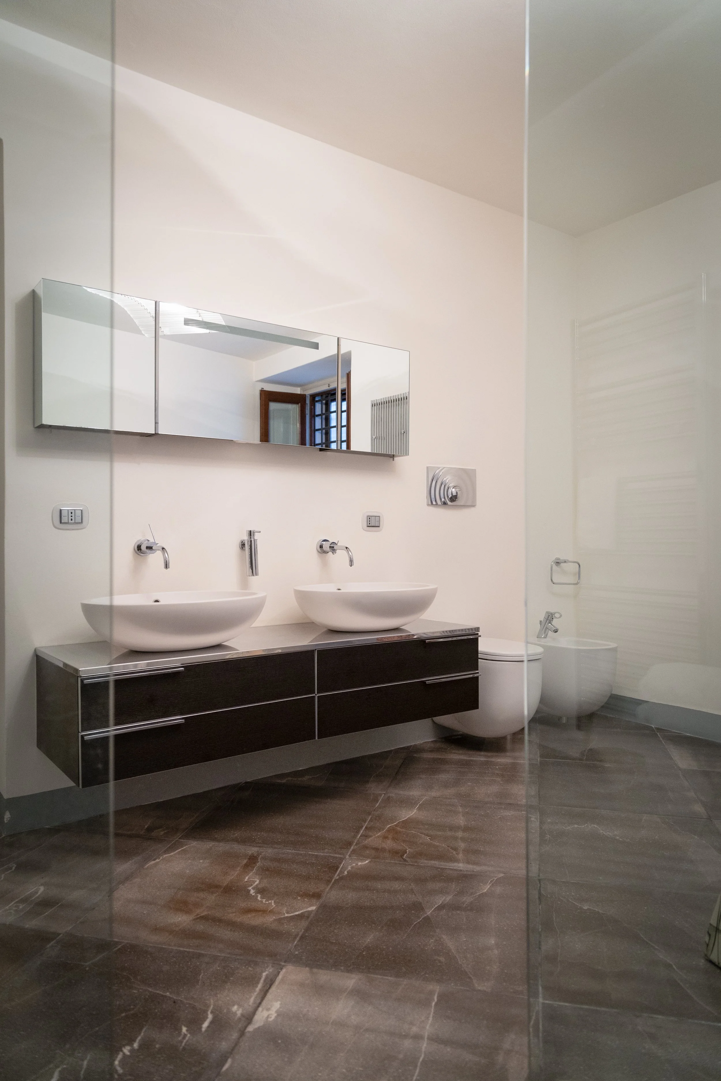 Modern bathroom with two white vessel sinks, large mirror, dark floating vanity, and a toilet and bidet. Tiled floors and a glass partition.